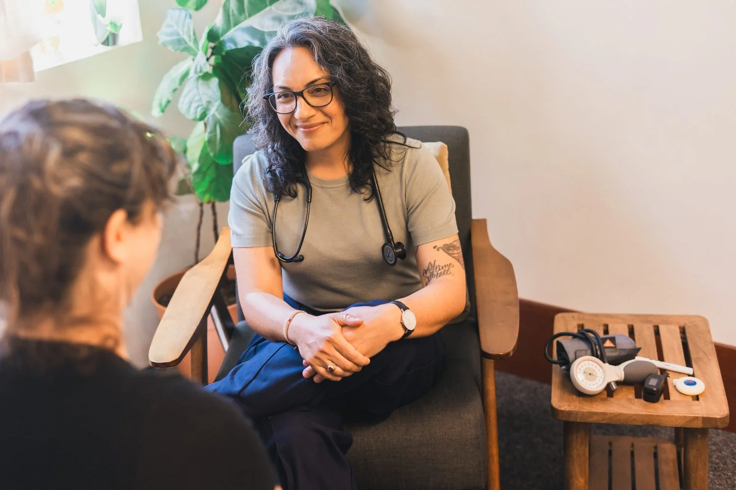 A female doctor with curly hair, glasses, and tattoos, smiling while talking to a patient in a consultation room. She has a stethoscope around her neck and is sitting on a chair. There is a wooden side table with medical equipment next to her.