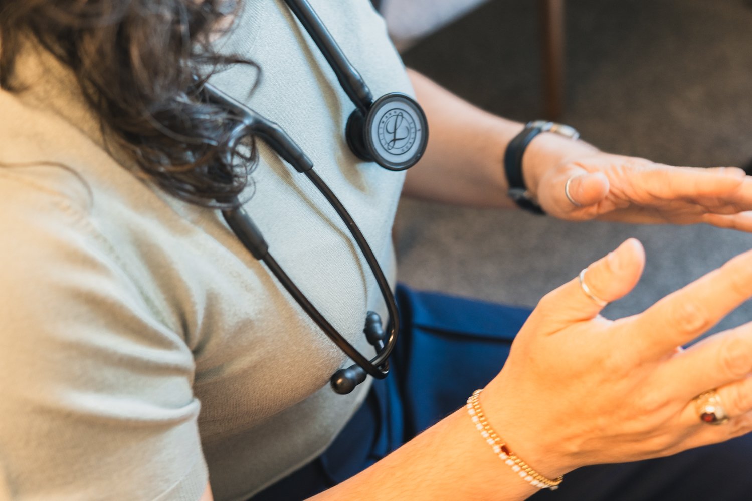 Close-up of a healthcare professional wearing a stethoscope around her neck, gesturing with her hands, wearing rings, bracelets, and a wristwatch.