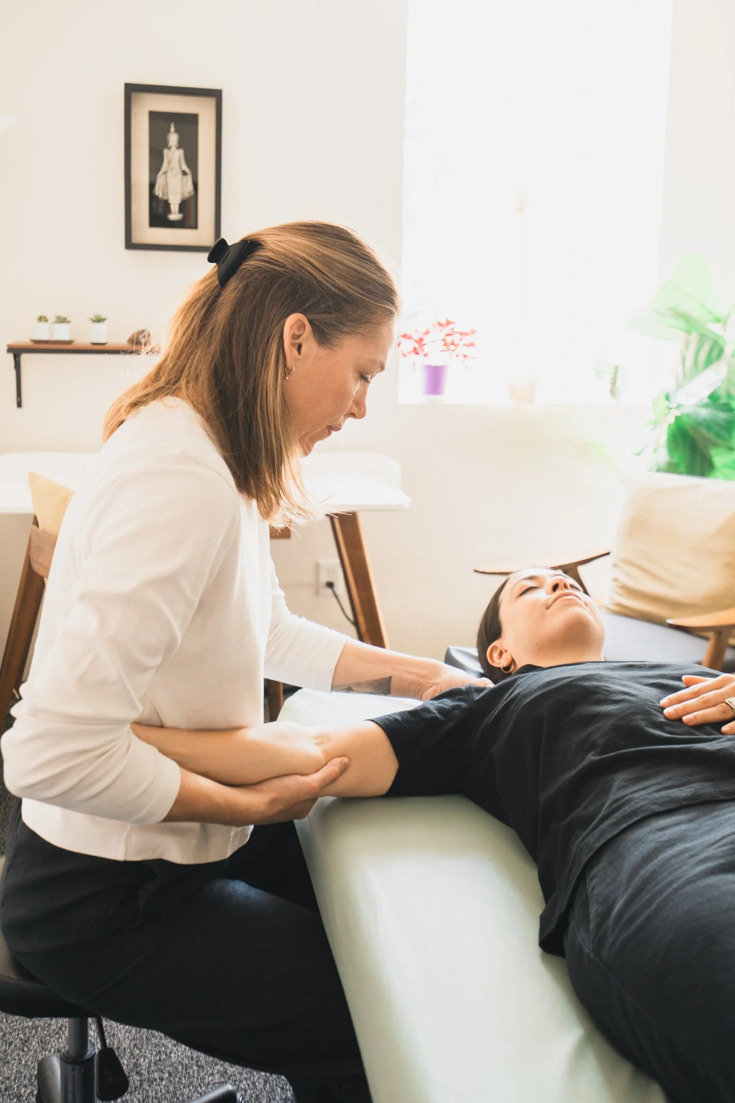 A woman lies on a table receiving physical therapy, with another woman assisting her by holding her arm and shoulder; the room has natural lighting, plants on the windowsill, and a framed picture on the wall.