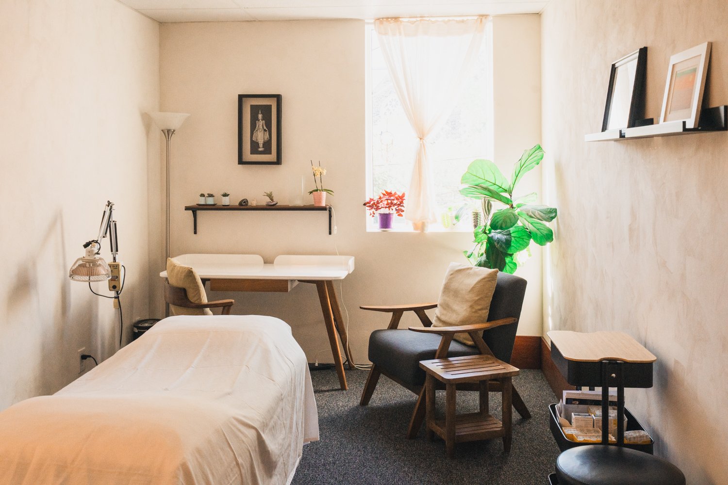 Small, cozy room with a bed, a black armchair, a wooden side table, a desk with chairs, potted plants near a window, framed pictures on the wall, and a shelf with small decorations.