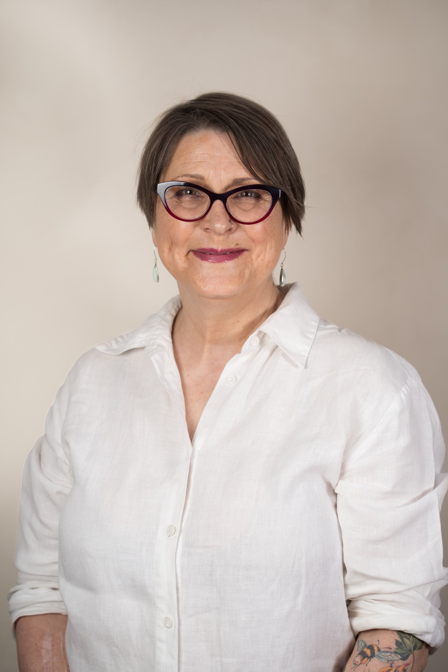 A woman with short dark hair, wearing glasses and a white shirt, smiling against a plain background.