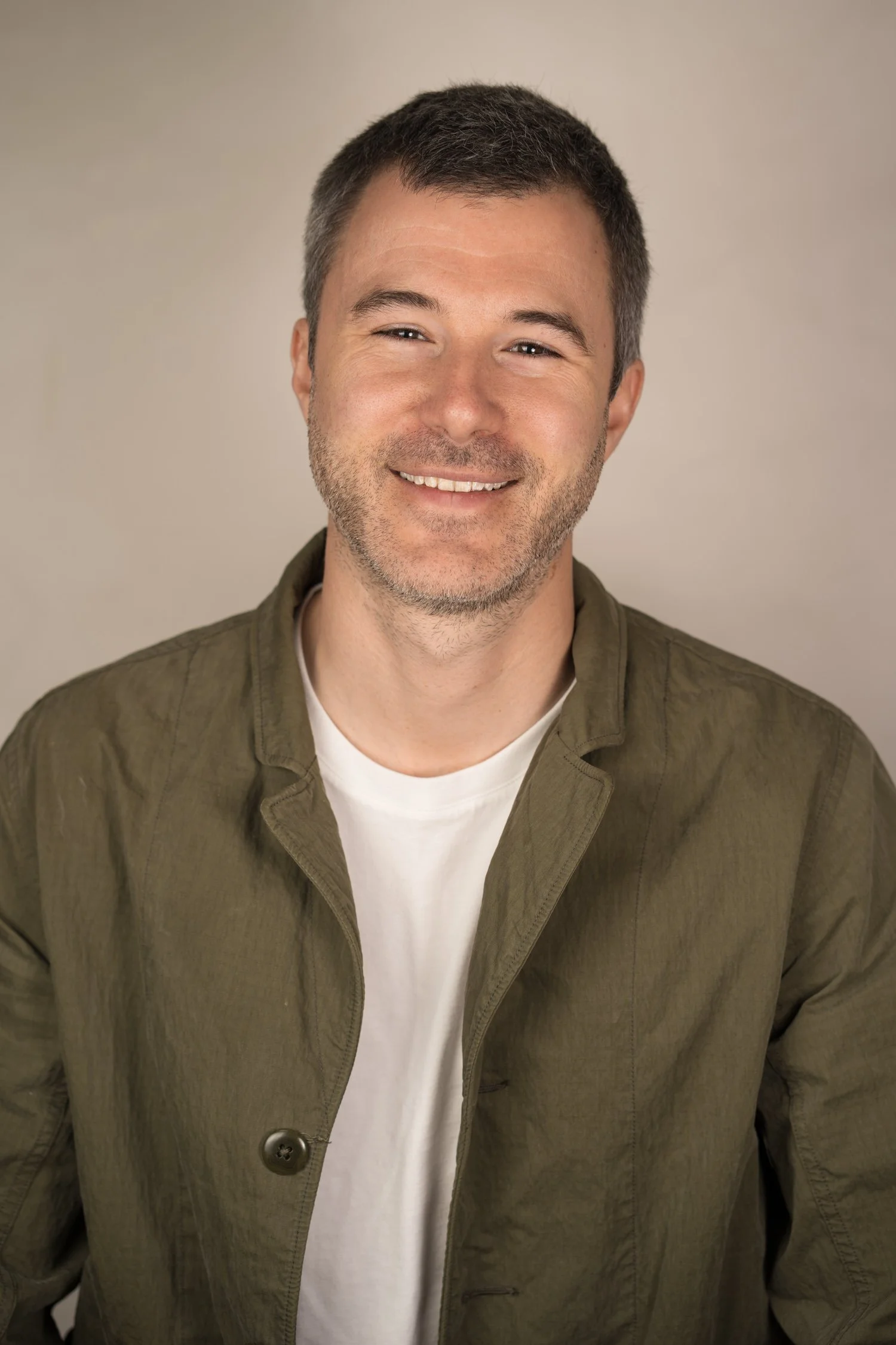 A smiling man with short dark hair and a beard, wearing an olive green jacket and a white T-shirt, against a plain beige background.