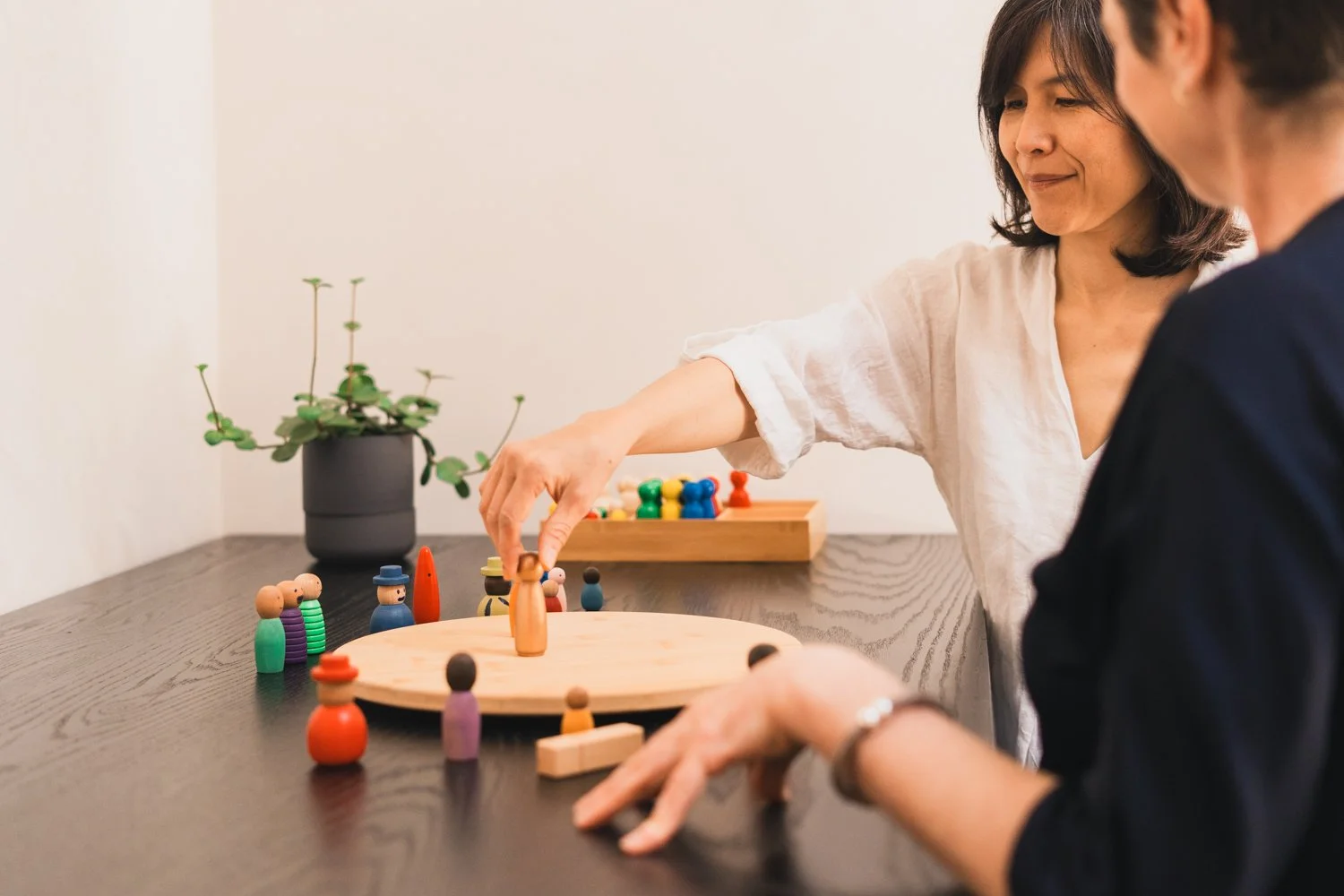 Two women playing a game with colorful wooden pegs on a round wooden board at a table, with a plant on the side.