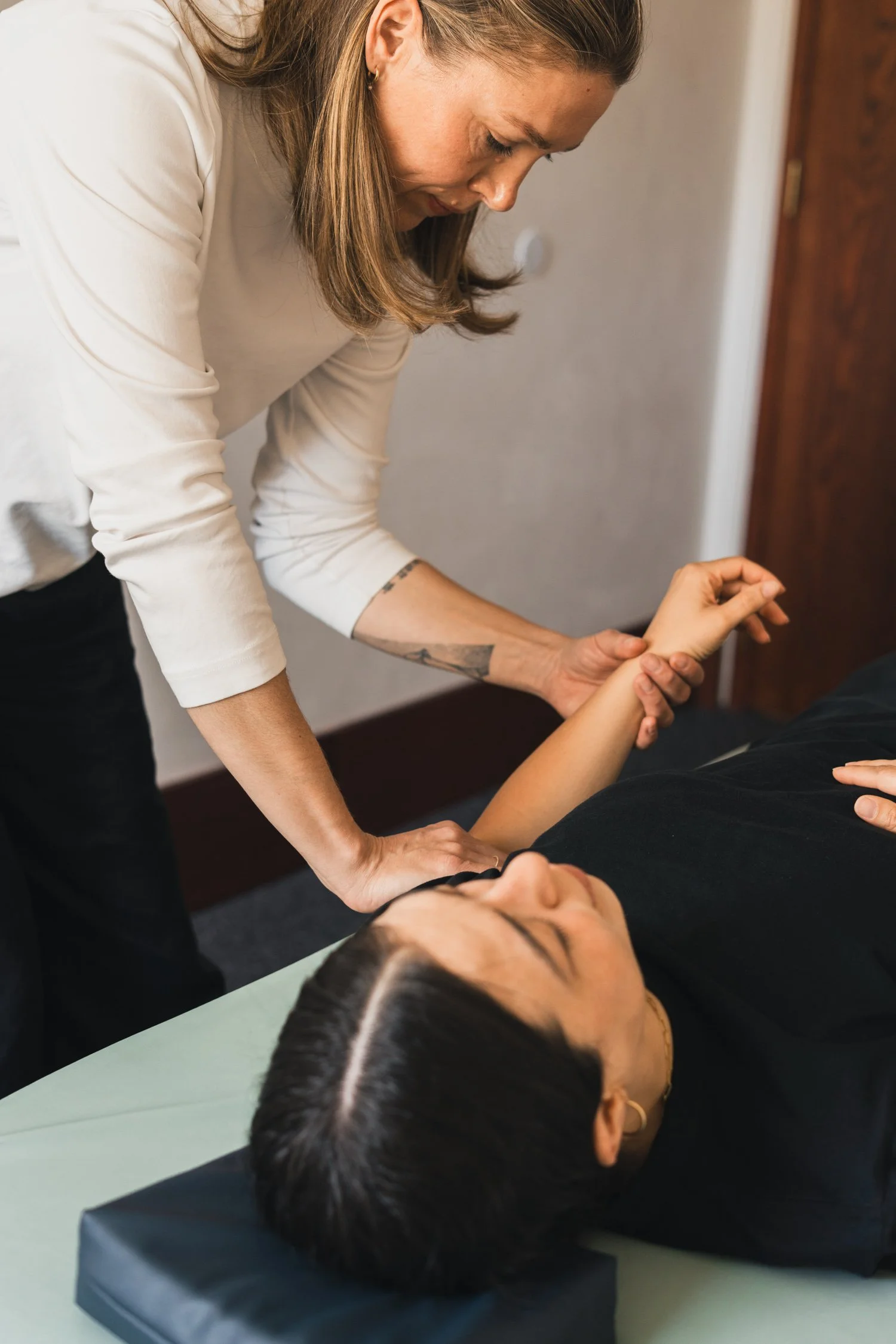 A woman receiving a massage from a massage therapist in a wellness clinic, lying on her back on a massage table with her eyes closed.