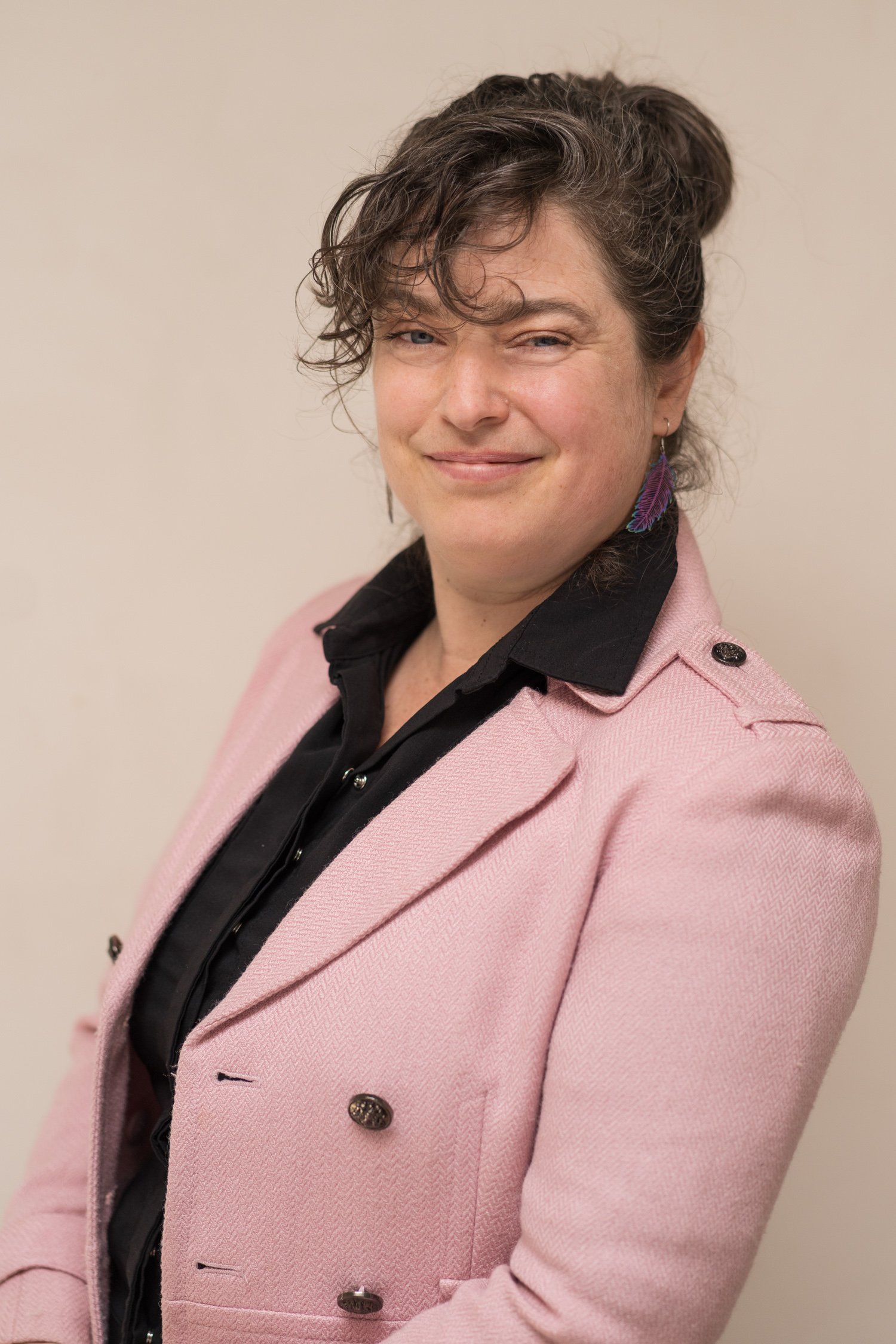 Portrait of a woman with curly brown hair and blue eyes, wearing a pink blazer, black shirt, and purple feather earring, standing against a plain beige background.