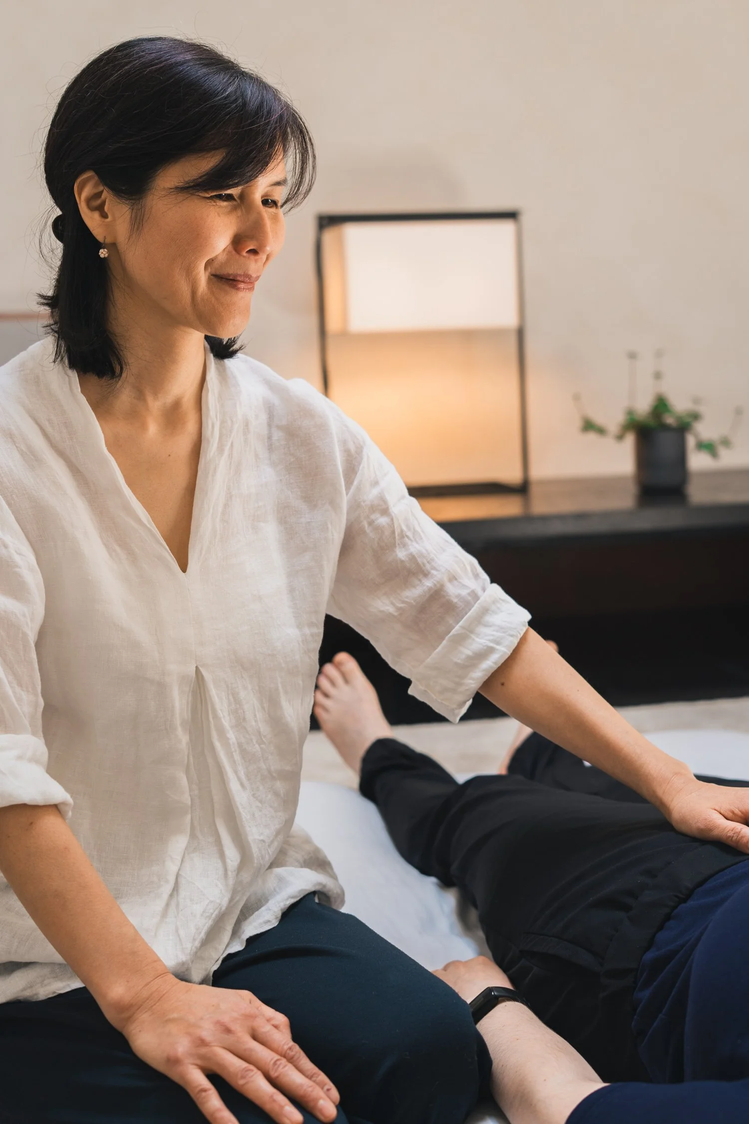 A woman with short dark hair dressed in a white blouse is giving a massage to a person lying on a massage table. The person receiving the massage is wearing black pants and a gray shirt and is lying face up with one arm resting on the table. The setting is a room with a lamp and a potted plant in the background.