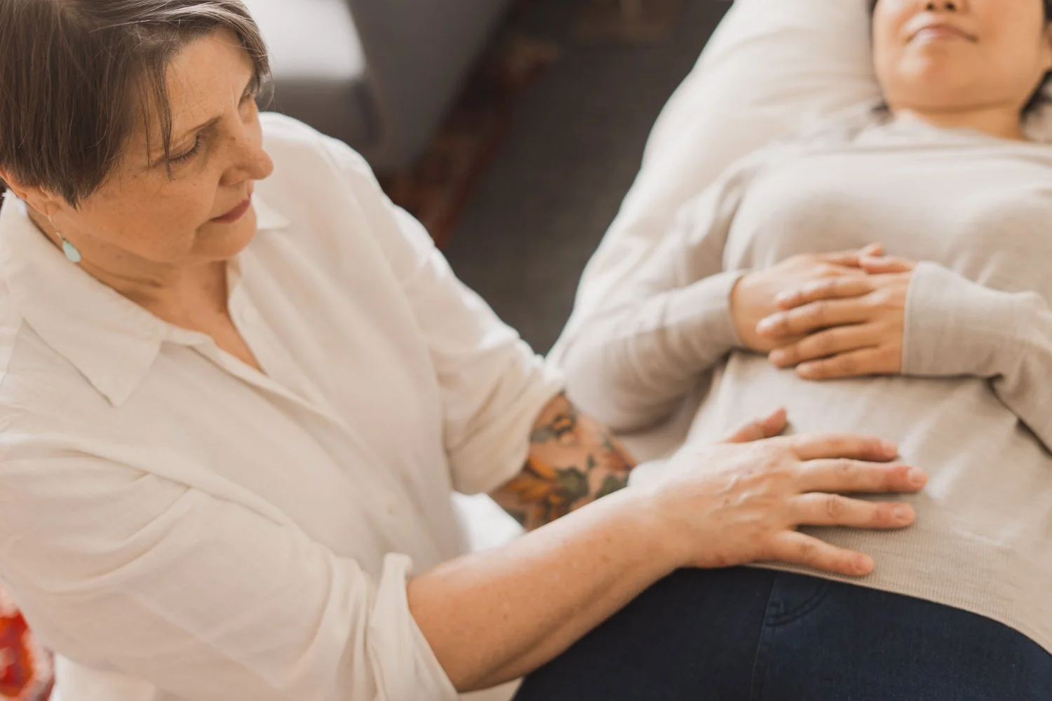 A woman performing a hands-on healing session or therapy on a person lying down with their hands resting on their stomach.