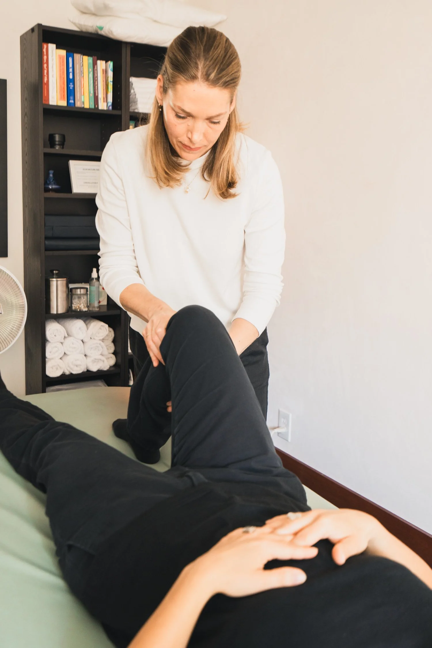 A woman in a white top and earrings is performing a physical therapy or massage on a person's leg, who is lying on a treatment table. In the background, there is a black bookshelf with books, towels, and therapy supplies.