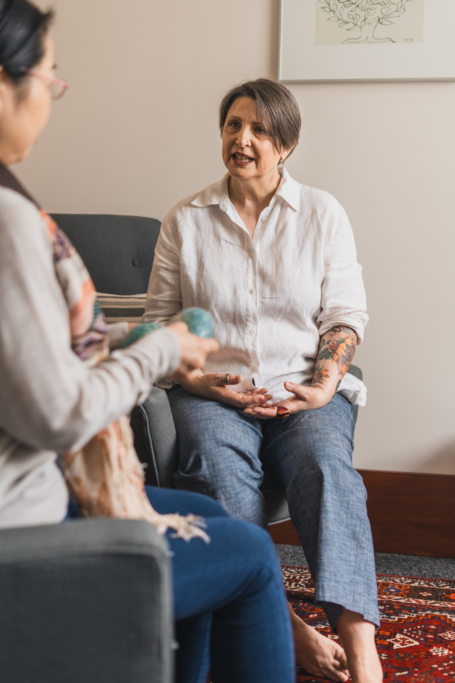 Two women engaged in a conversation in a cozy living room. One woman with short brown hair and tattoos on her arms is seated on a chair, wearing a white shirt and jeans, speaking and using hand gestures. The other woman, partially visible with dark h