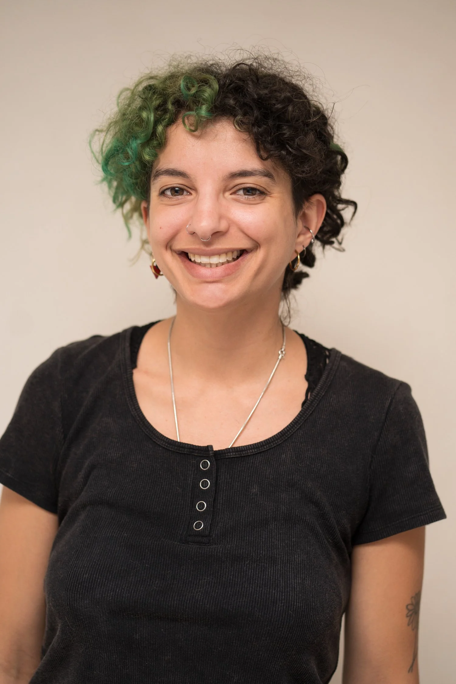 A young woman with short, curly hair featuring green highlights, smiling, wearing a black top, earrings, a nose ring, and a necklace, standing against a plain background.
