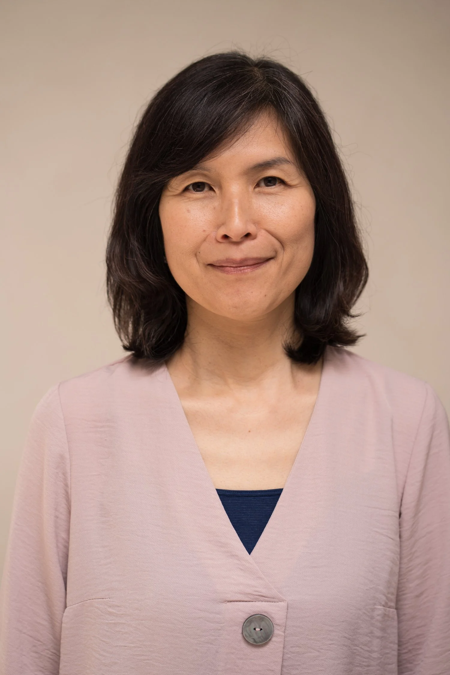 A woman with shoulder-length dark hair, wearing a pink blazer with a large button and a dark top underneath, standing against a plain, cream-colored background.