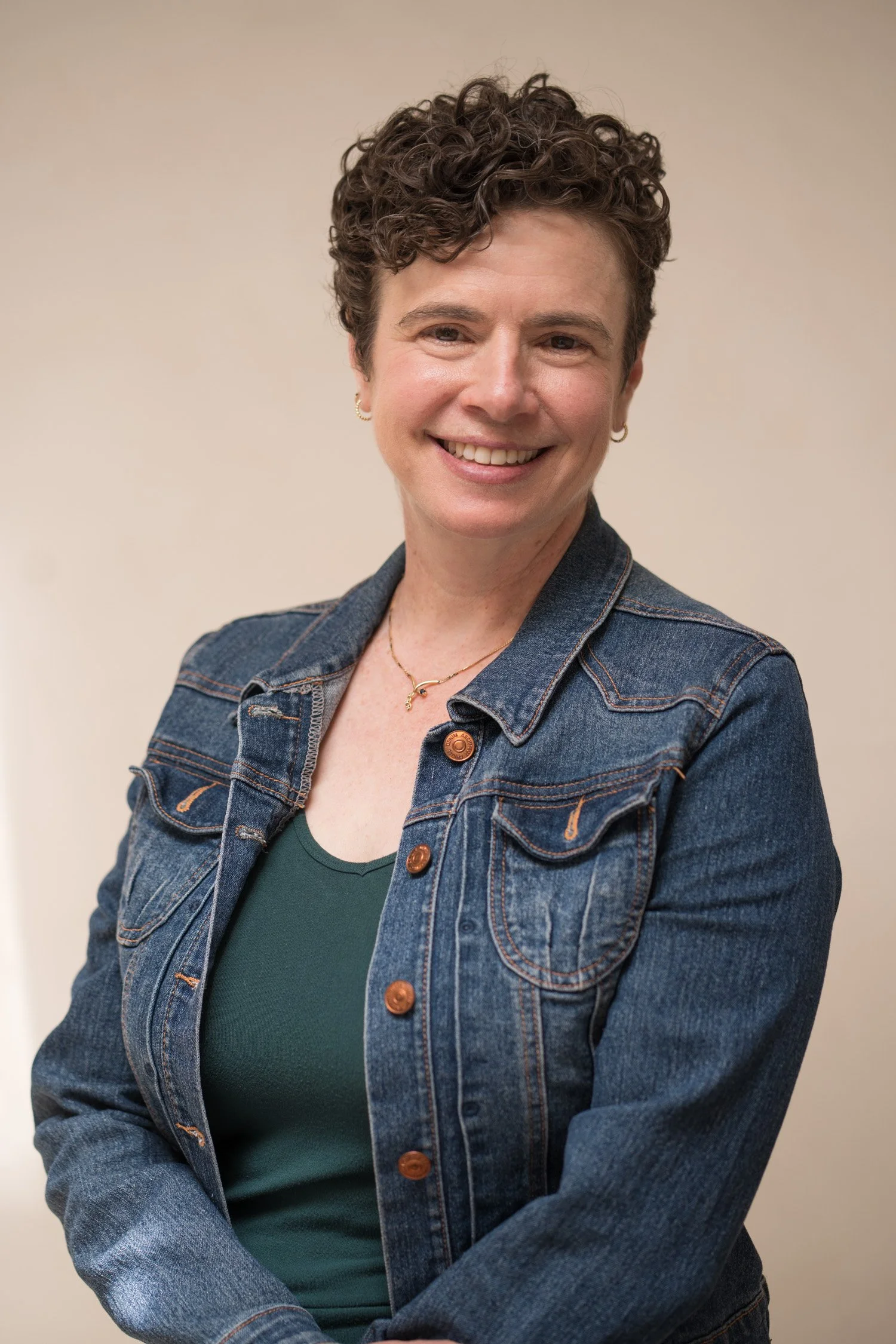 A woman with short, curly hair smiling and wearing a denim jacket and a dark green top, against a plain beige background.