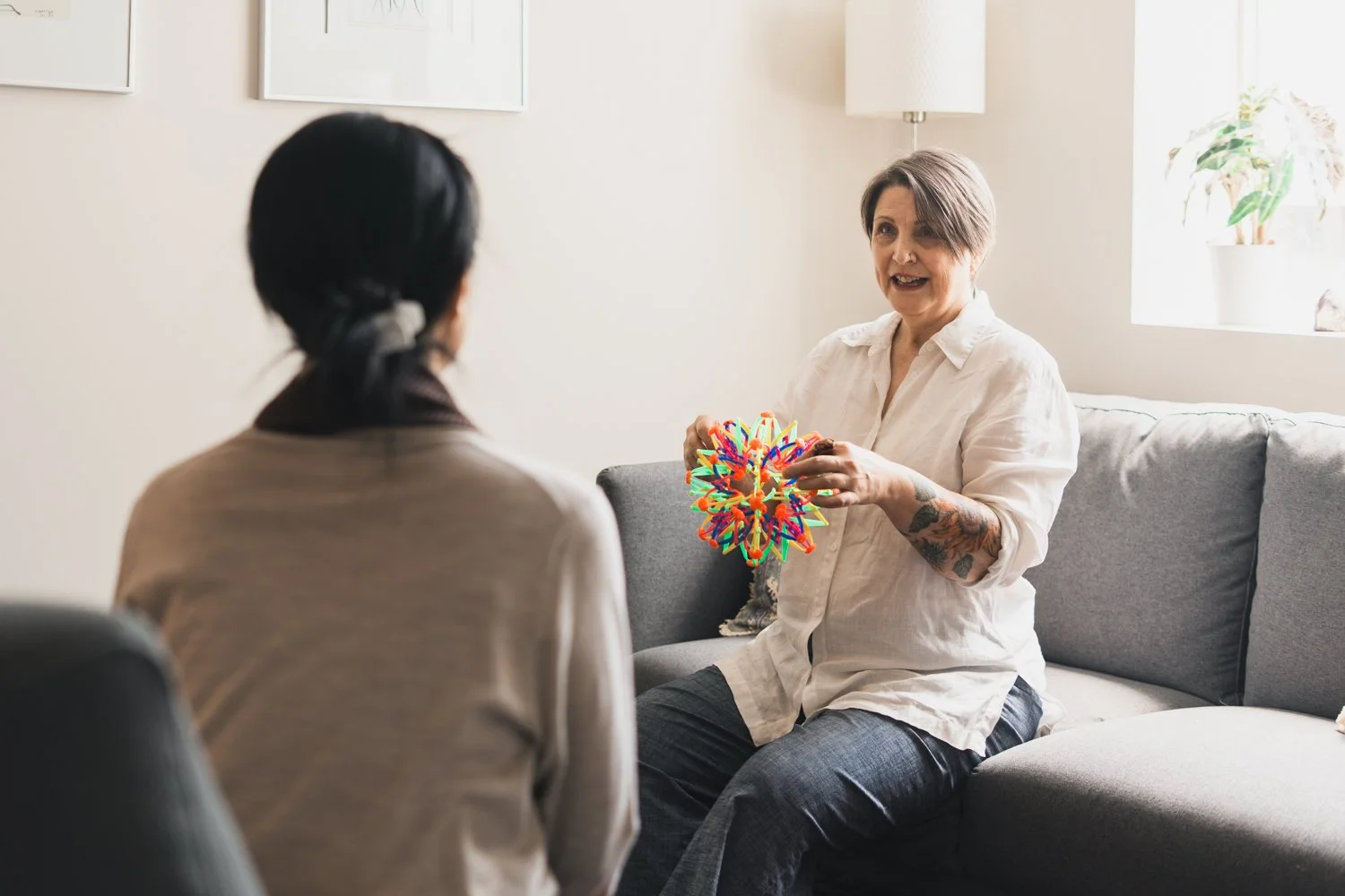 A woman with tattoos on her arm is sitting on a sofa, holding a colorful, spiky puzzle toy, and talking to another woman with dark hair, facing away from the camera, in a cozy living room with a window and framed artwork.