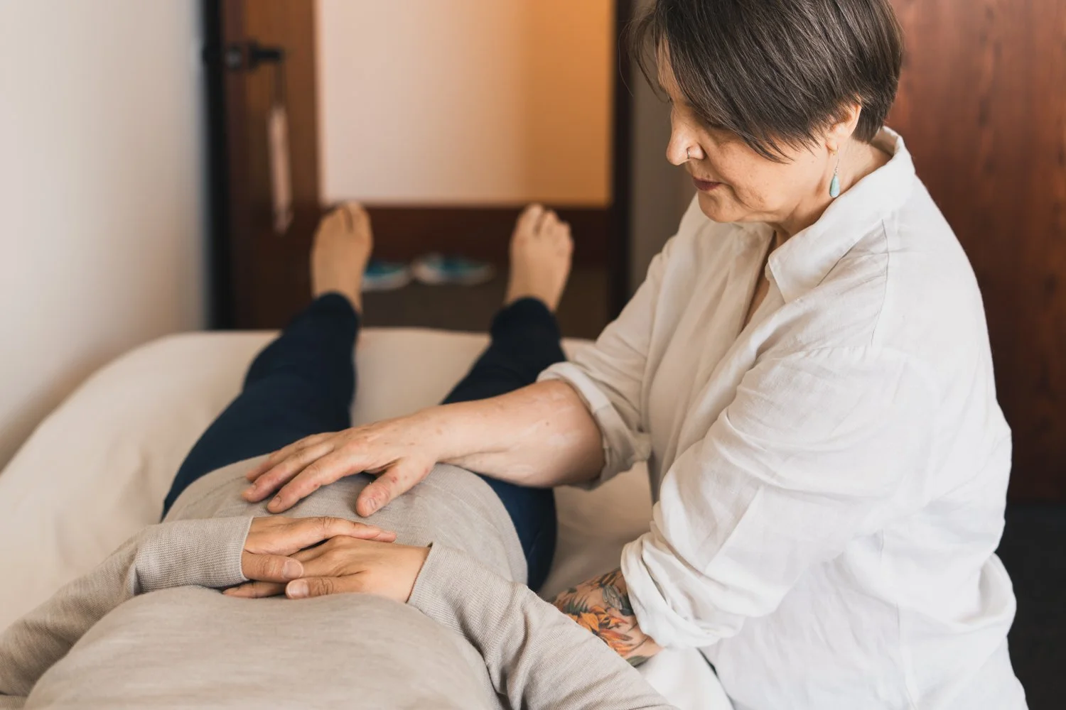 A woman receiving a massage or therapy on her lower back, lying on a therapy table, with her hands resting on her stomach, while the therapist, wearing a white shirt, works on her back in a room with warm lighting.