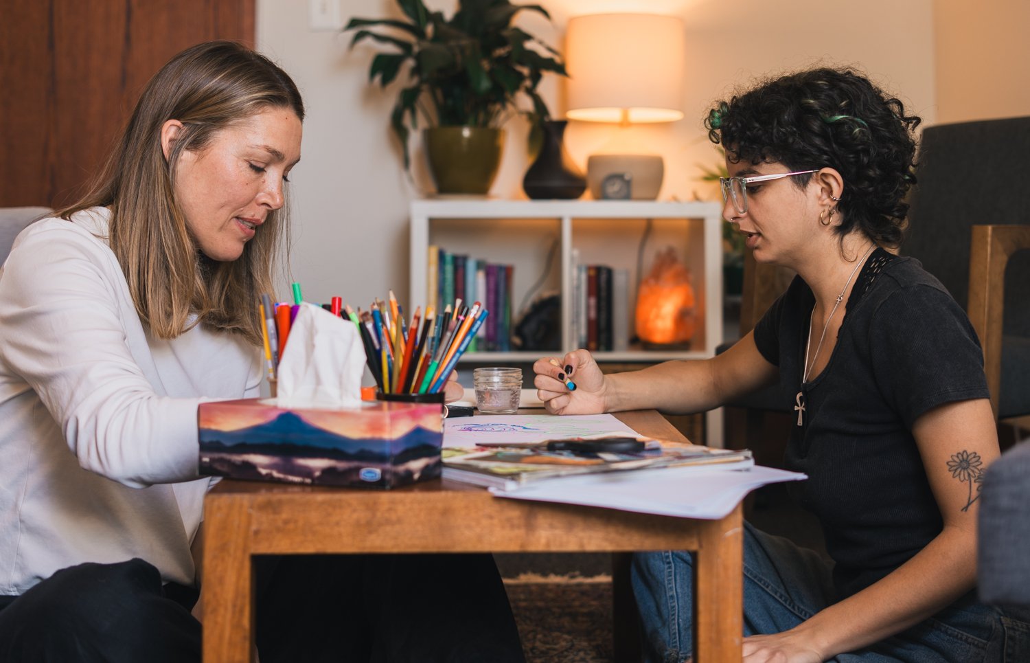 Two women sitting at a wooden table engaged in a conversation or activity, with art supplies and papers in front of them, in a cozy room with bookshelves and warm lighting.