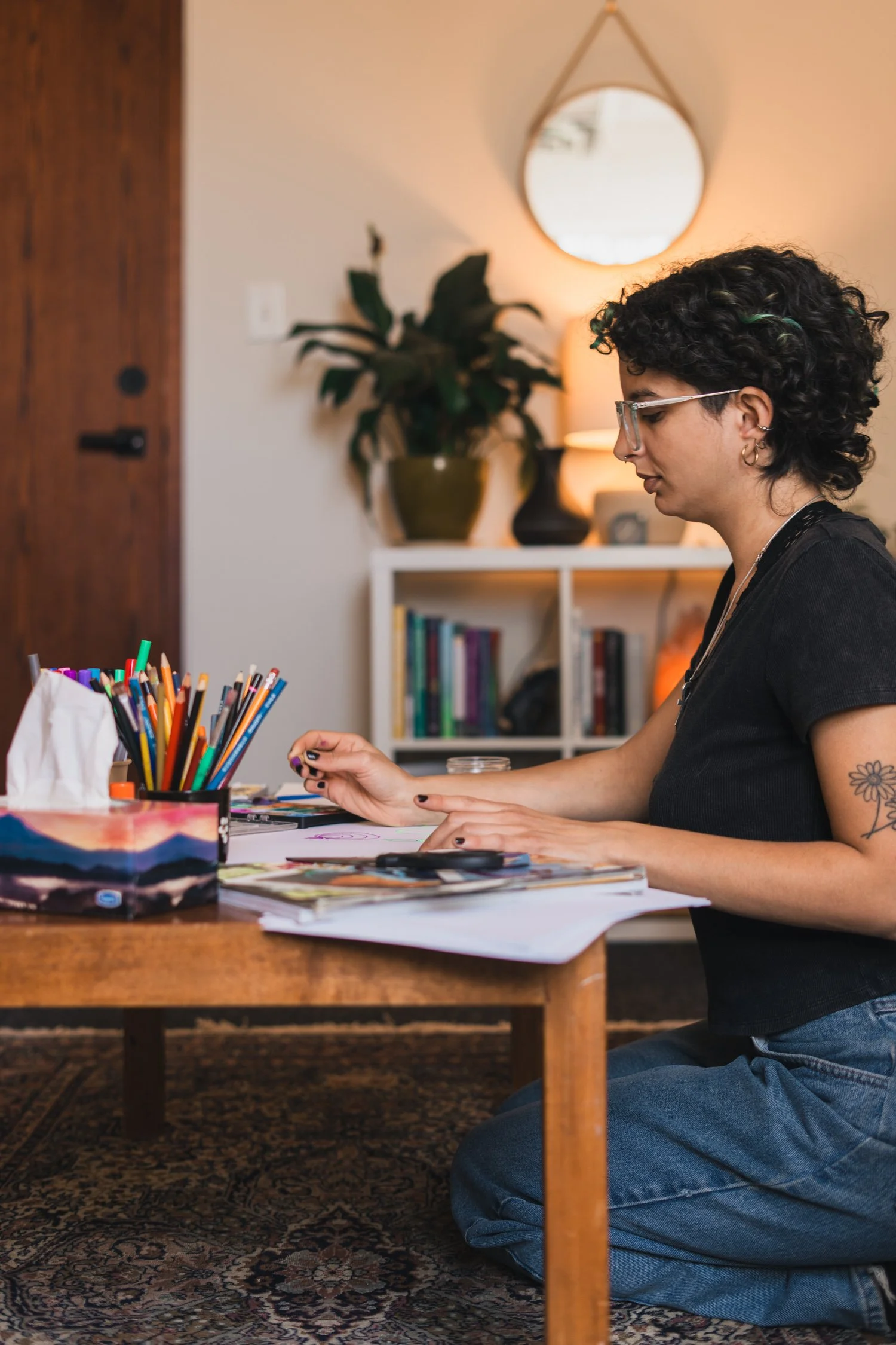A woman with short curly hair, glasses, and tattoos, sitting on the floor at a wooden table, engaging with art supplies, with a decorated living room in the background.