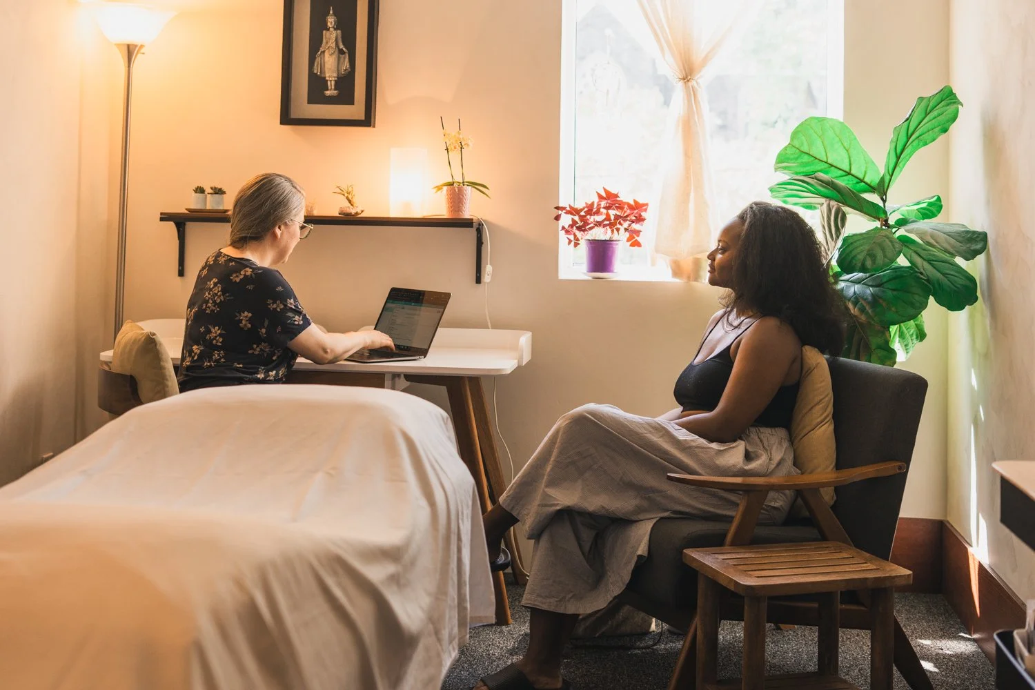 Two women having a consultation in a cozy room with plants, a window, and soft lighting.