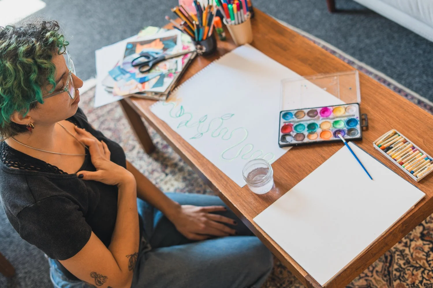 A person sitting at a wooden table with art supplies, including watercolor paints, brushes, markers, and a blank sheet of paper, preparing to paint, with a glass of water nearby.