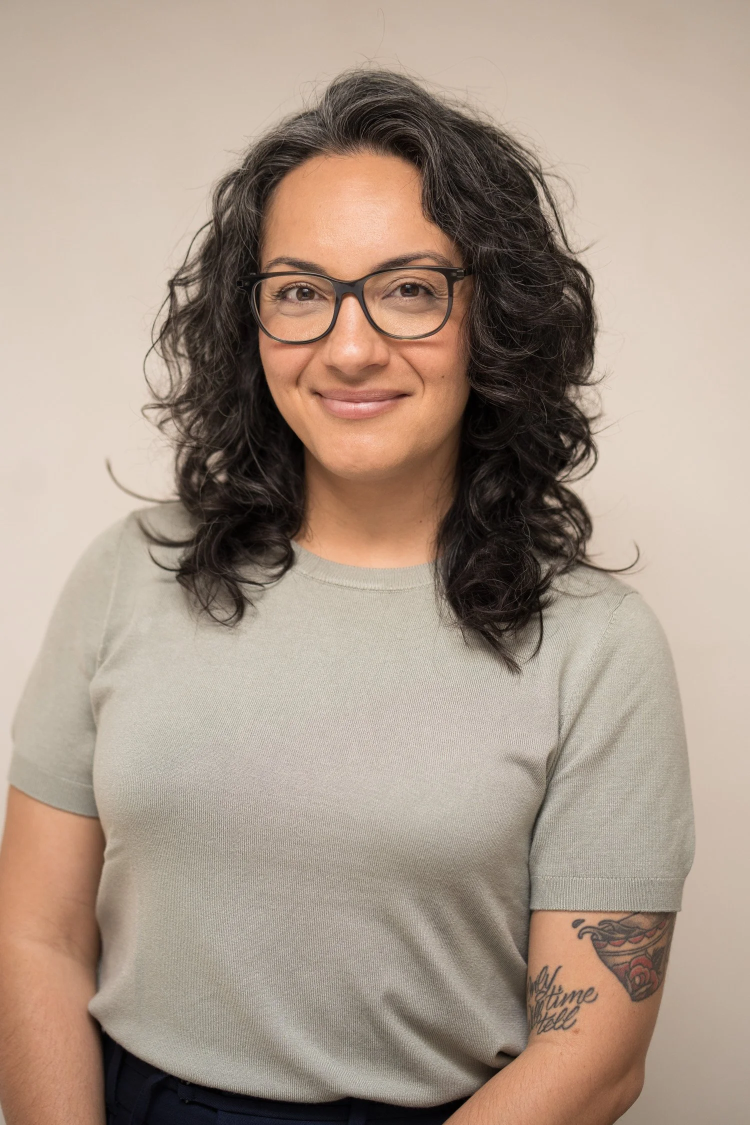 Close-up portrait of a woman with curly dark hair wearing glasses and a light gray t-shirt, smiling softly against a plain beige background.