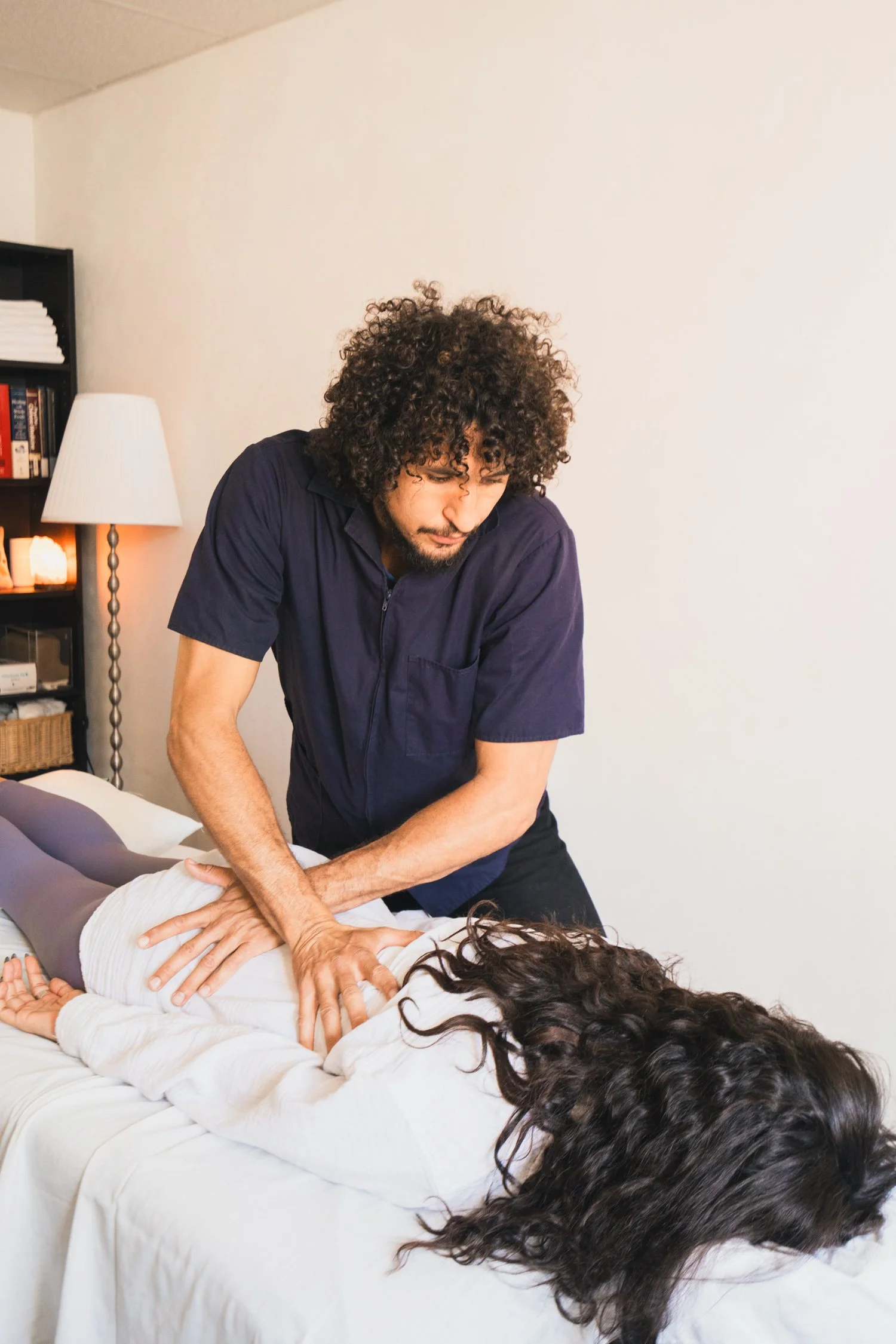 A person performing CPR on a patient lying on a bed, with a caregiver observing in a room with a lamp and bookshelf.