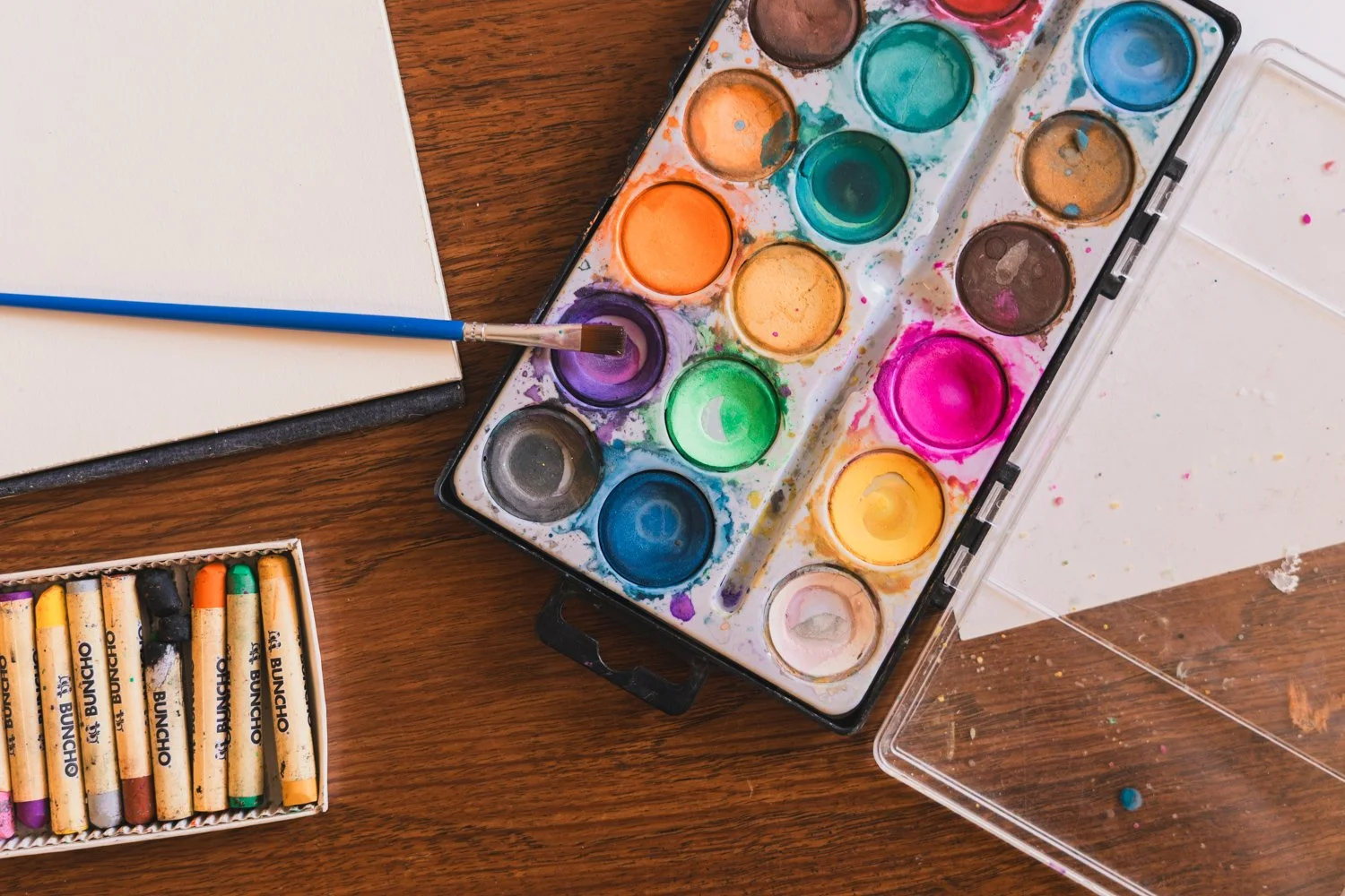 An open watercolor paint set with various colors, a paintbrush resting on a purple paint, a box of crayons, a blank notebook, and a white sheet of paper on a wooden table.