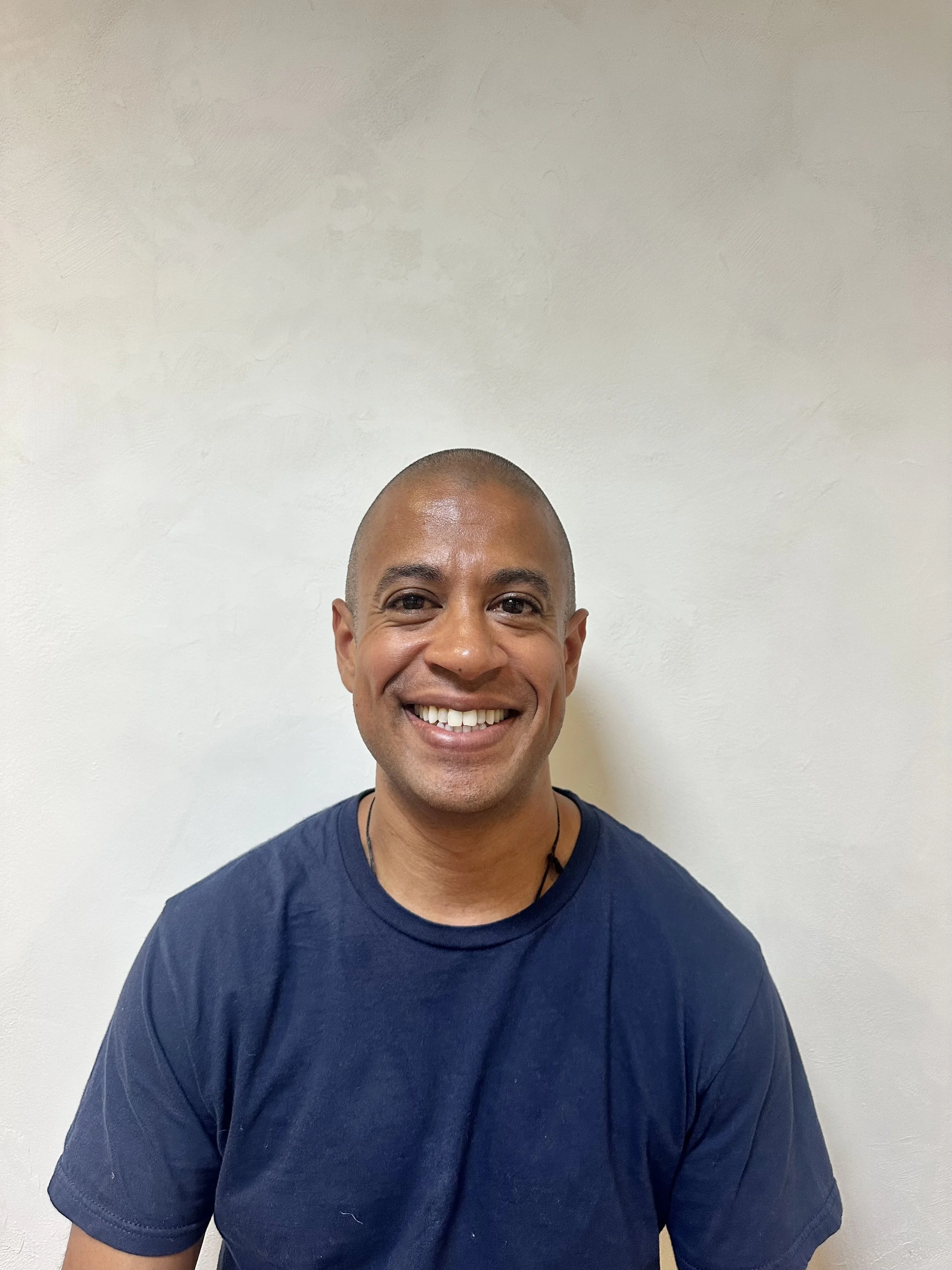 Smiling man with a shaved head and wearing a navy blue T-shirt, standing against a plain light-colored wall.