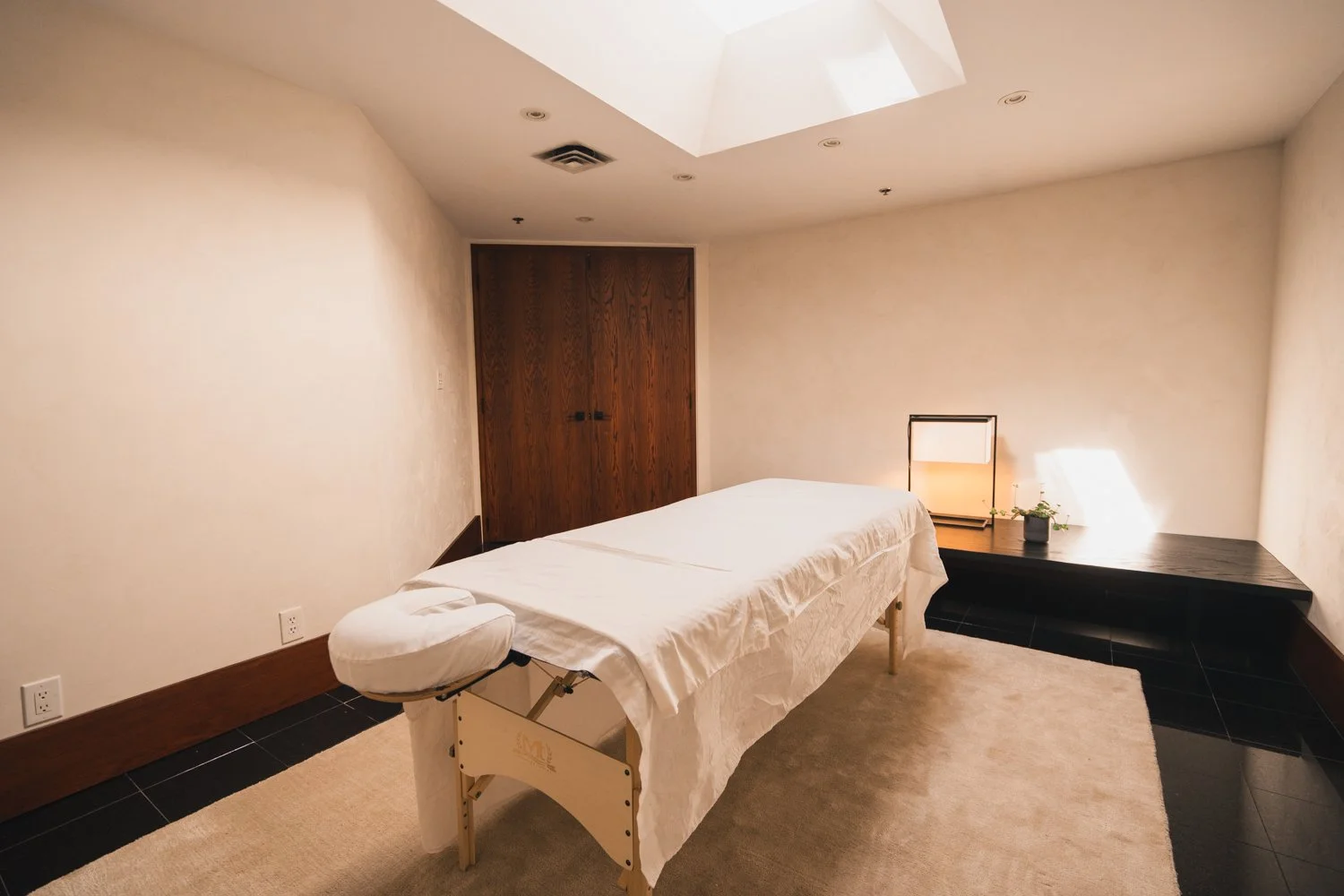 A massage room with a massage table covered in a white sheet, a small black side table with a lamp and a plant, a beige carpet, beige walls, a wooden door, a skylight, and recessed ceiling lights.