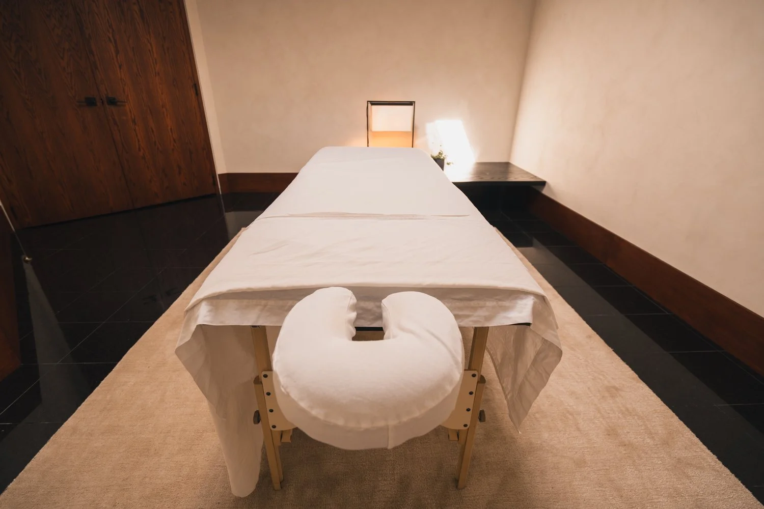 A massage table with a white sheet and a U-shaped pillow in a softly lit room with beige walls, black tile flooring, and a wooden door.
