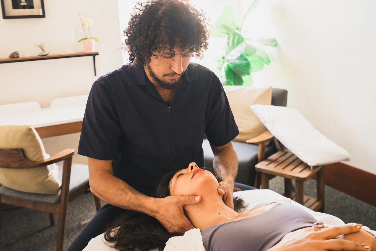 A man providing a massage to a woman lying on a massage table in a cozy room with plants and chairs.