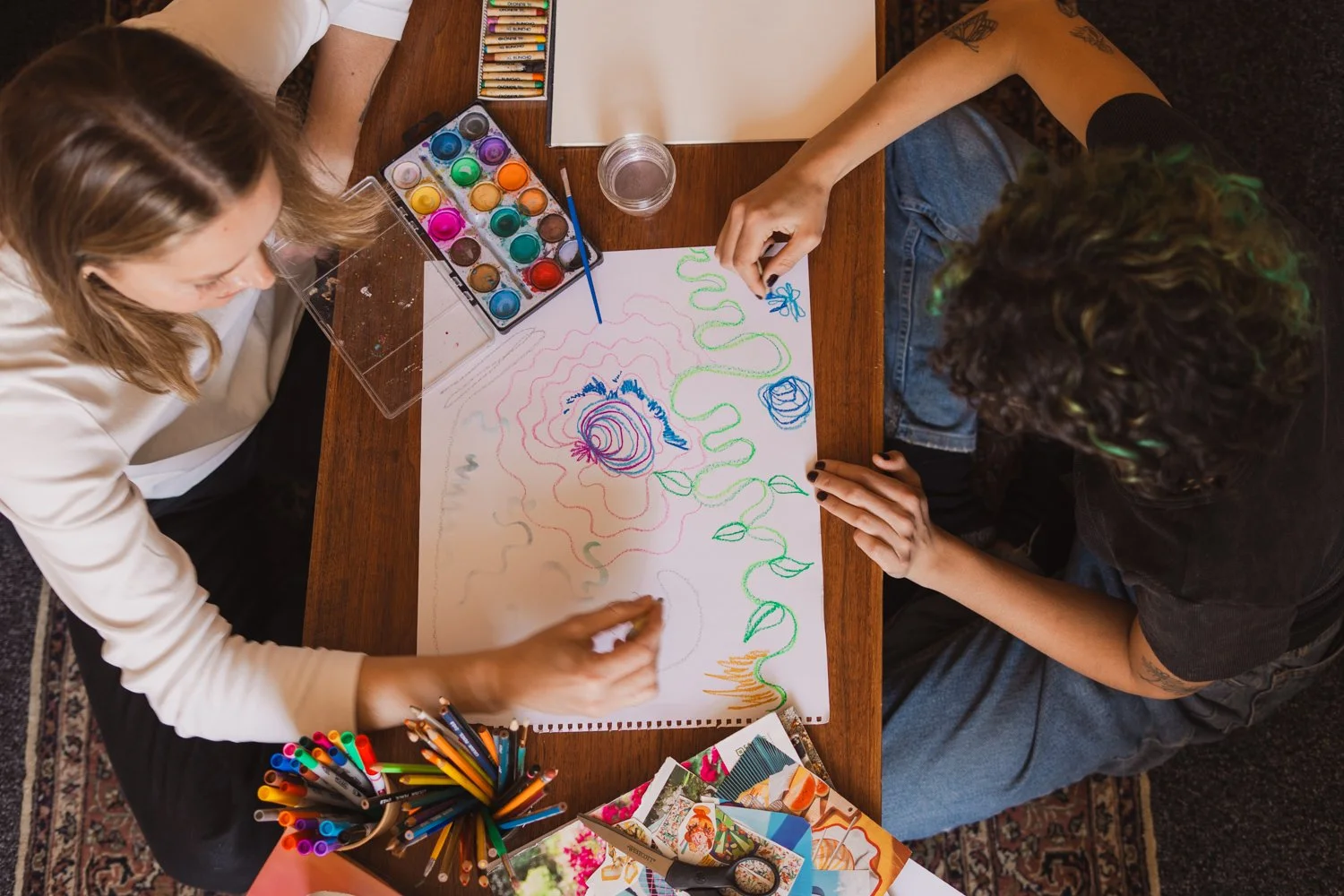 Two women are drawing and painting on a large sheet of paper on a wooden table, surrounded by art supplies including watercolor paints, markers, colored pencils, and photos.