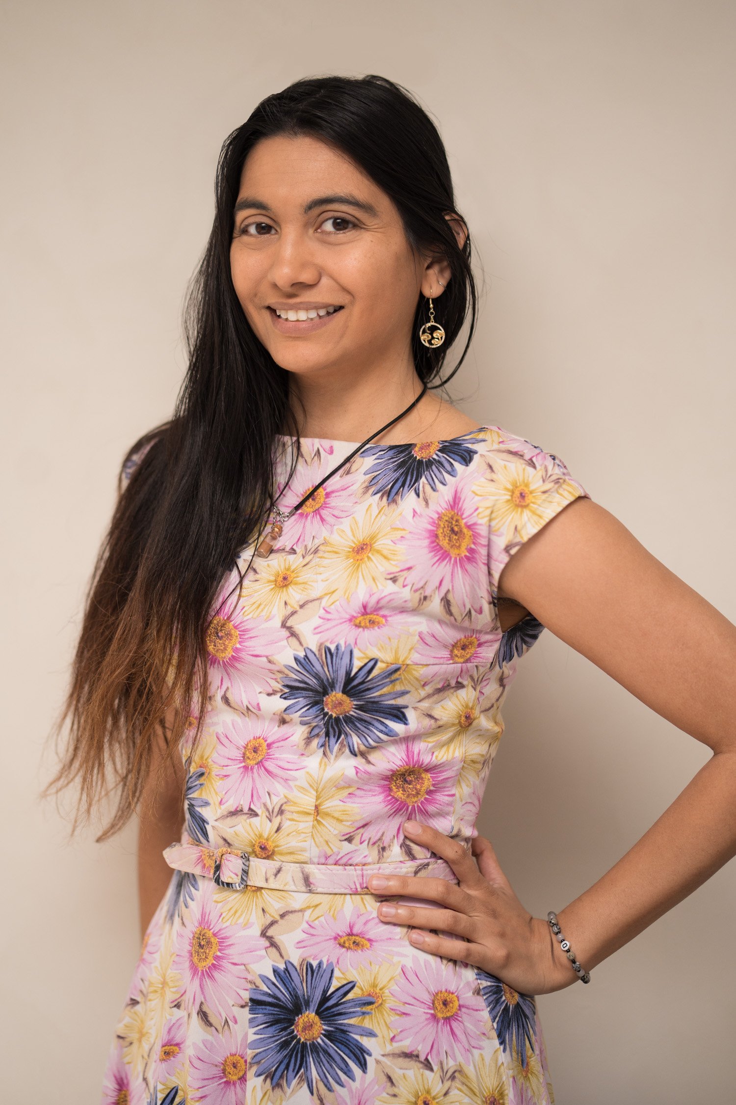 A woman with long dark hair, smiling, wearing a floral dress with pink, yellow, and blue flowers, accessorized with earrings, a necklace, a bracelet, and a ring, standing against a plain light-colored wall.