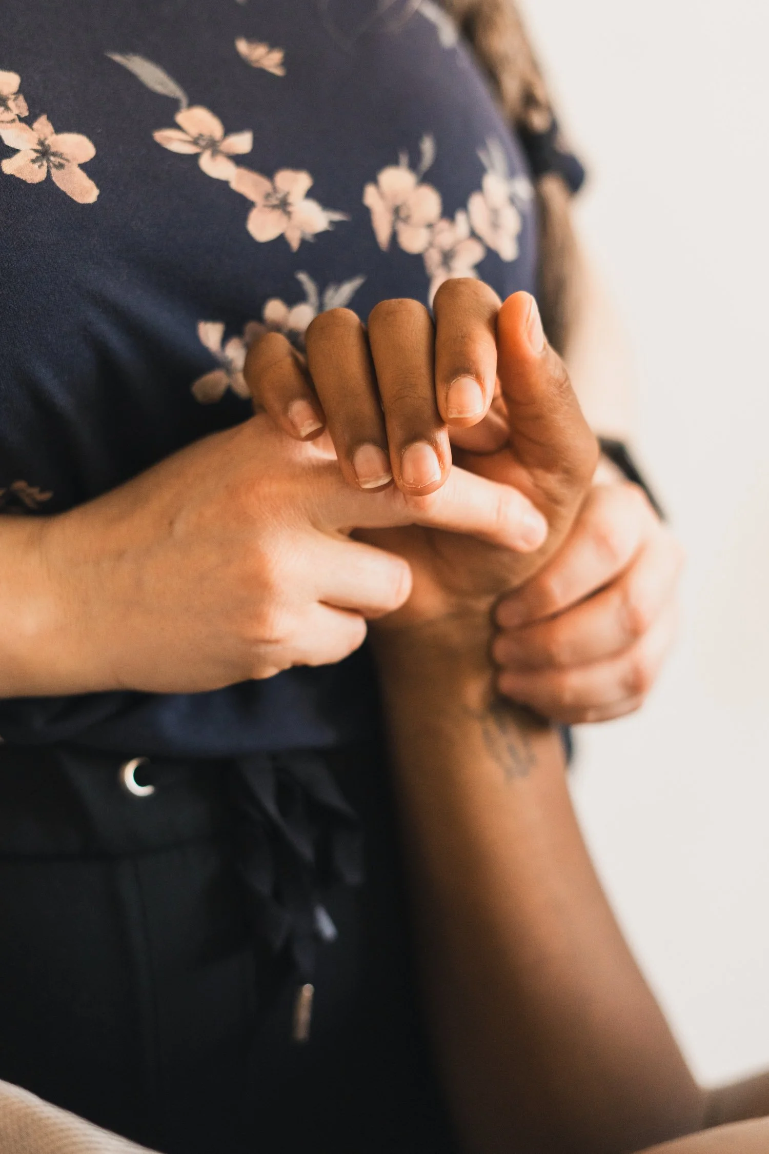 Close-up of two hands holding each other, one with darker skin and the other with lighter skin, against a background of a person's torso wearing a dark shirt with a pink floral pattern.