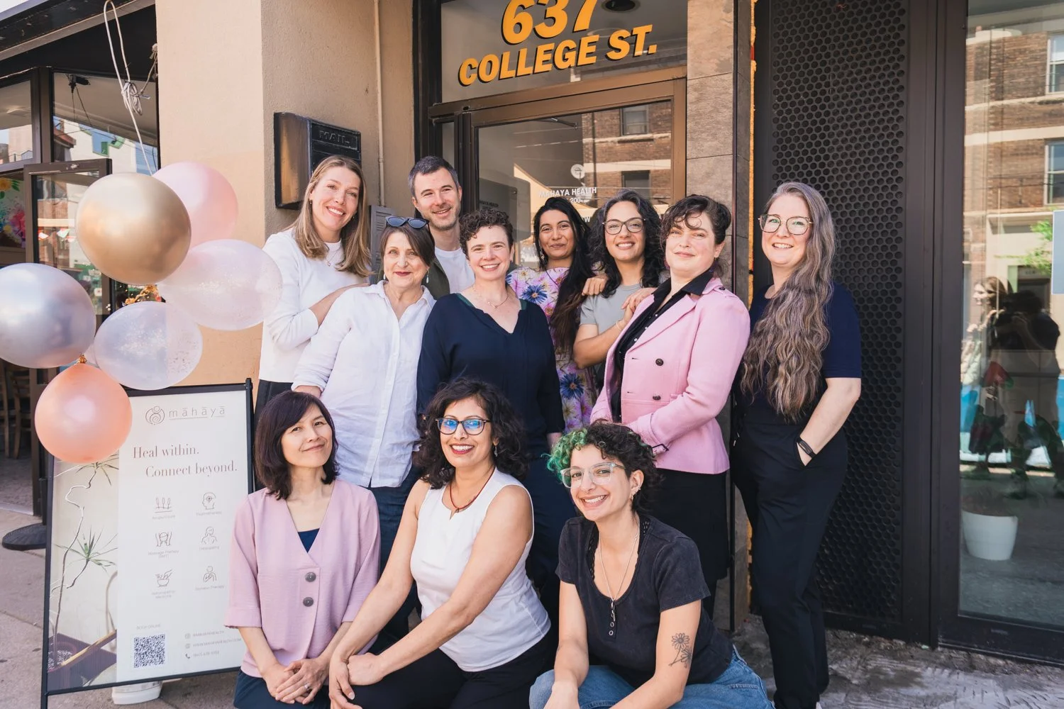 Group of eleven women standing and sitting outside a building with a sign reading '637 College St.' The women are smiling and dressed in casual to business casual clothing. There are pink, white, and gold balloons, and a sign with a QR code and some text next to the group.