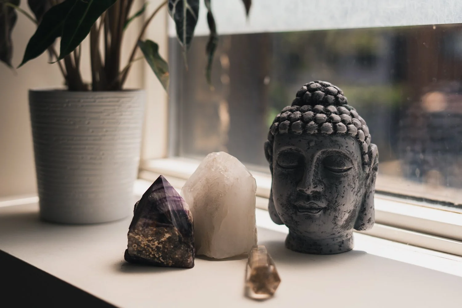 Decorative objects on a windowsill including a potted houseplant, a small purple and brown mineral, a white stone, a clear amber crystal, and a black and white stone Buddha head sculpture.