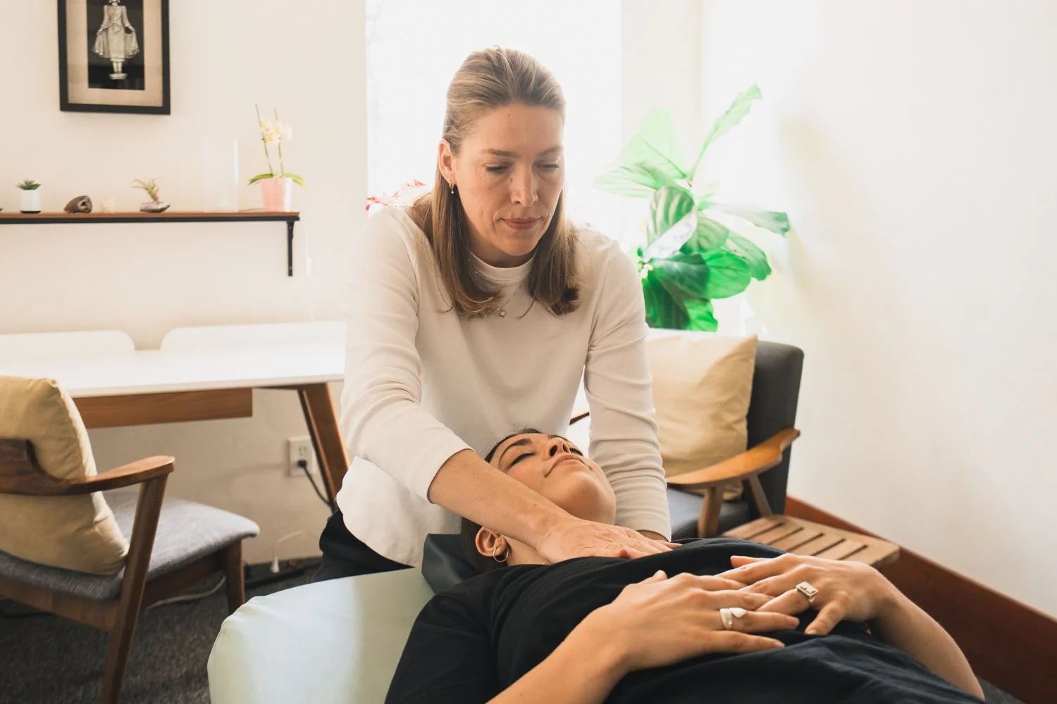 Woman receiving reiki massage from practitioner in a cozy, well-lit room with plants and decorative items.