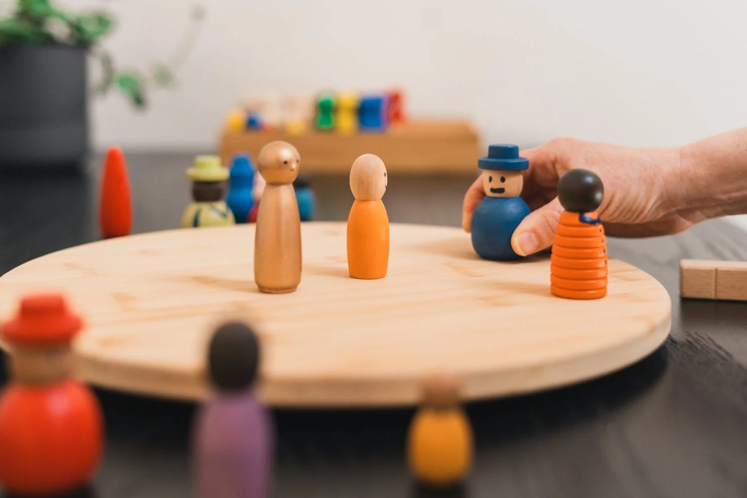 Close-up of wooden peg dolls used in a board game, placed on a round wooden board, with a person moving a blue peg doll wearing a hat. Other colorful peg dolls are visible in the background and foreground.