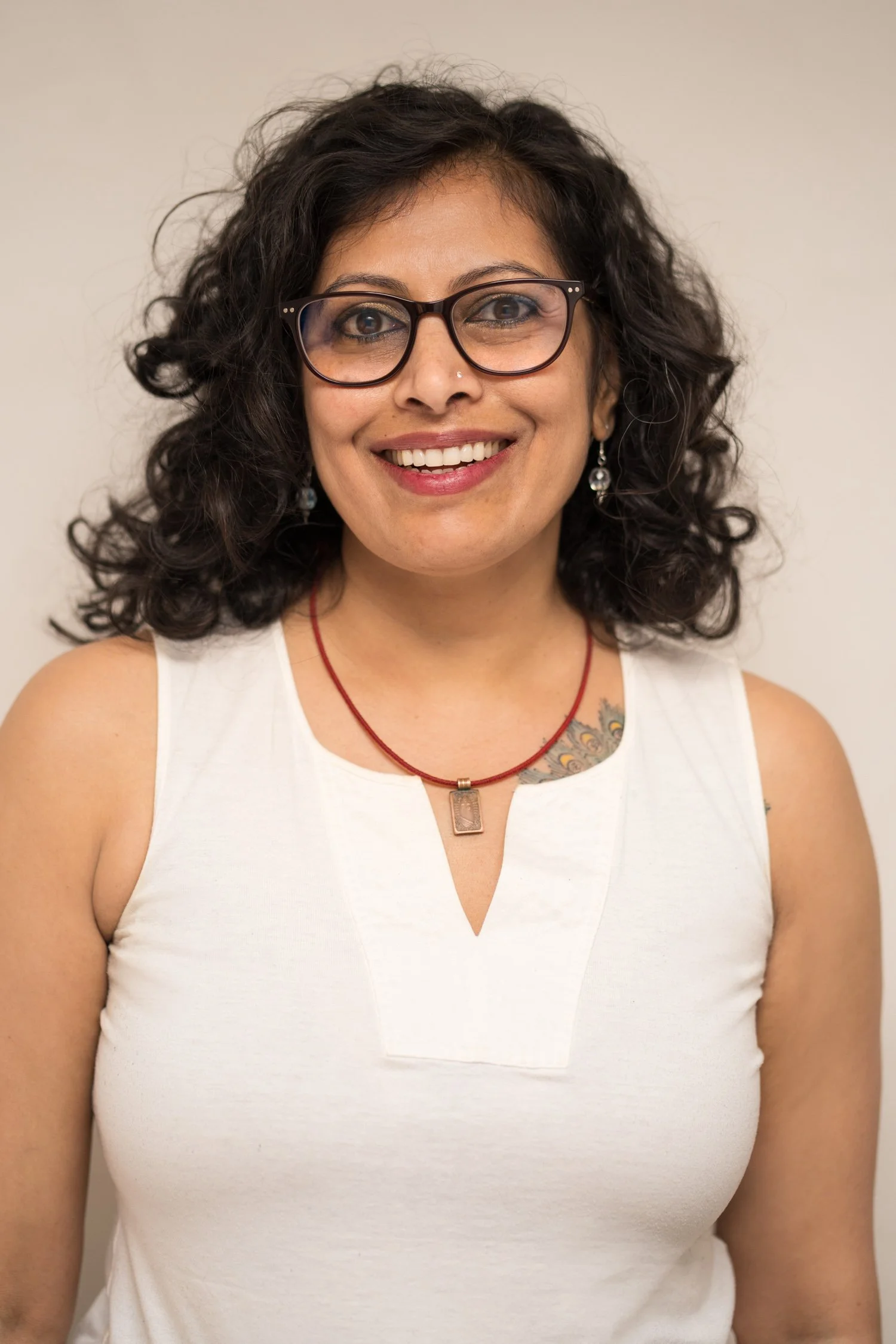 Close-up of a woman with curly dark hair wearing glasses, earrings, a white sleeveless top, and a red necklace, smiling against a plain background.