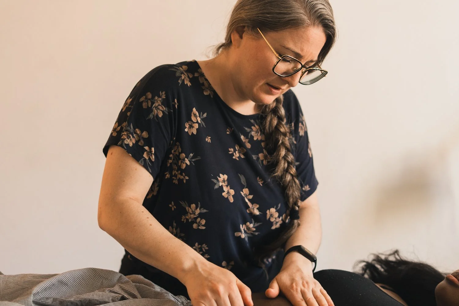 A woman with glasses and a floral t-shirt performs a back massage on a person lying face down on a massage table.