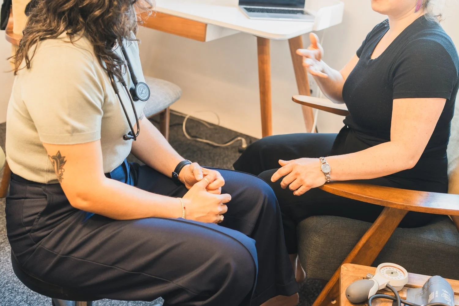 A healthcare professional wearing scrubs and a stethoscope consulting with a woman sitting in a chair, discussing medical information in an office setting.
