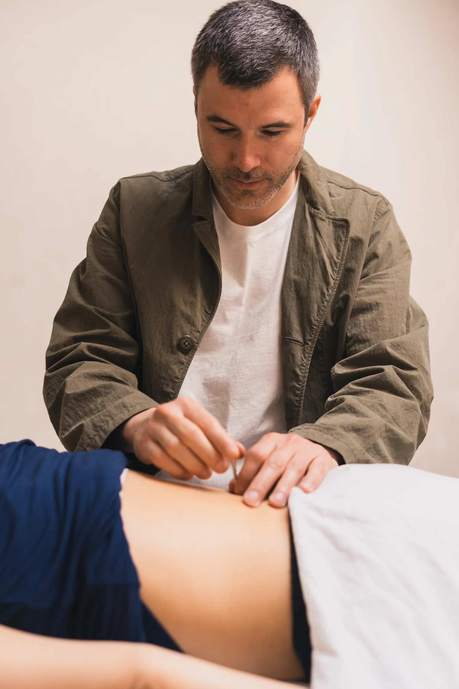 A man with gray hair and a beard, wearing a gray jacket and white shirt, performing CPR on a person lying on the ground, with another person wearing a dark blue shirt assisting.