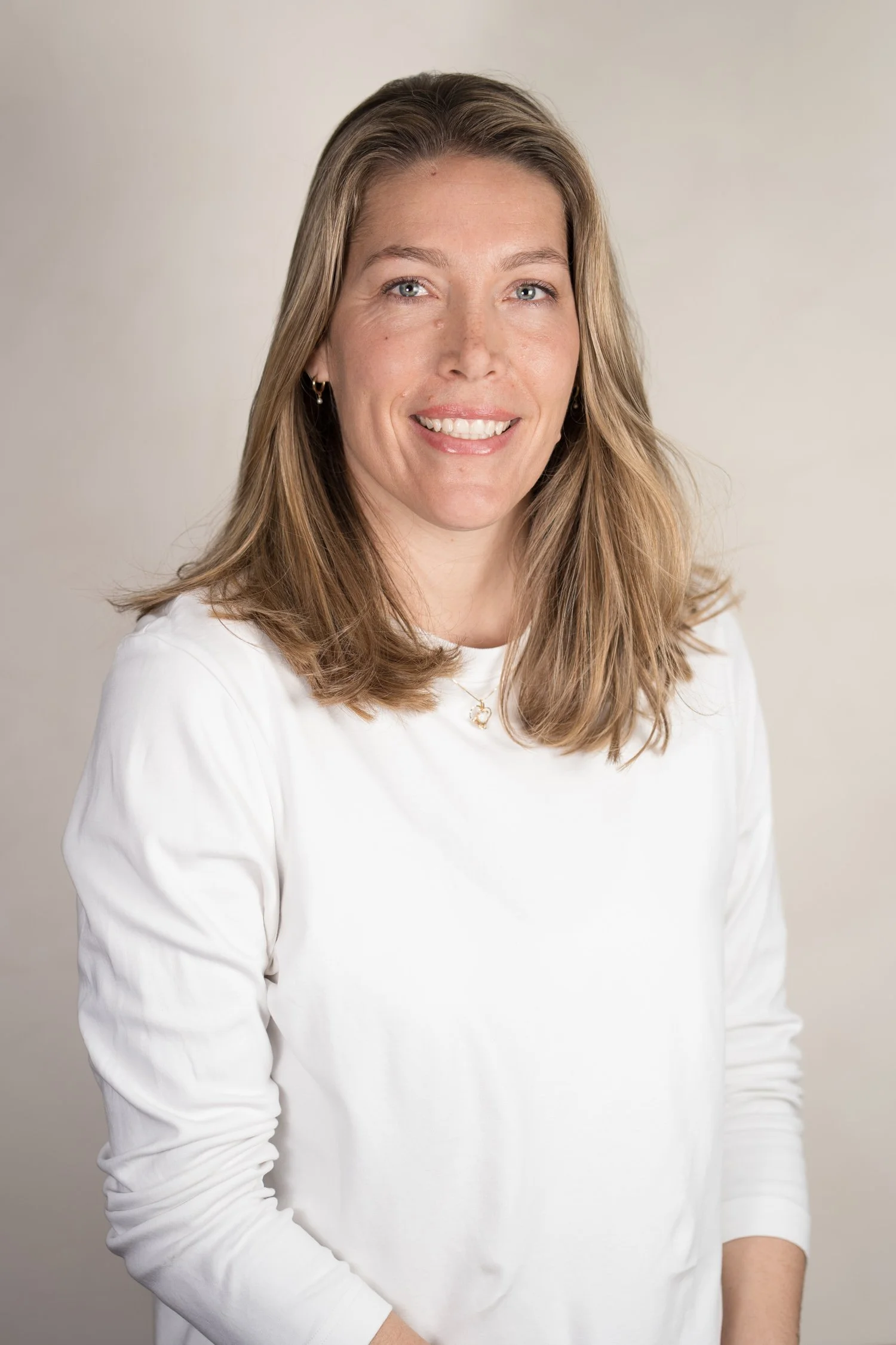A woman with light skin and medium-length wavy light brown hair, wearing a white long-sleeve shirt and small hoop earrings, smiling, against a neutral background.
