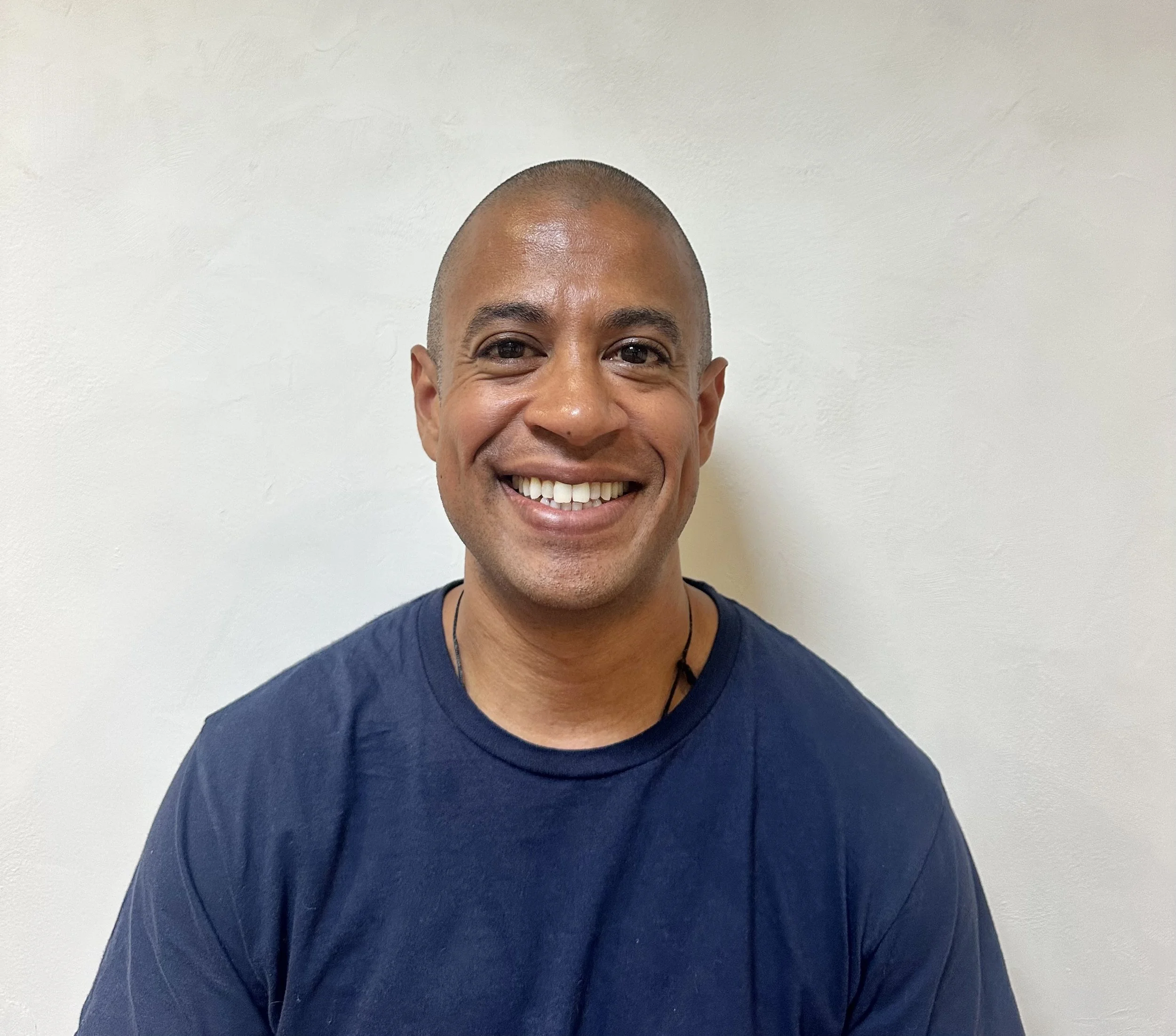 A smiling man with a shaved head wearing a navy blue shirt standing in front of a plain white wall.