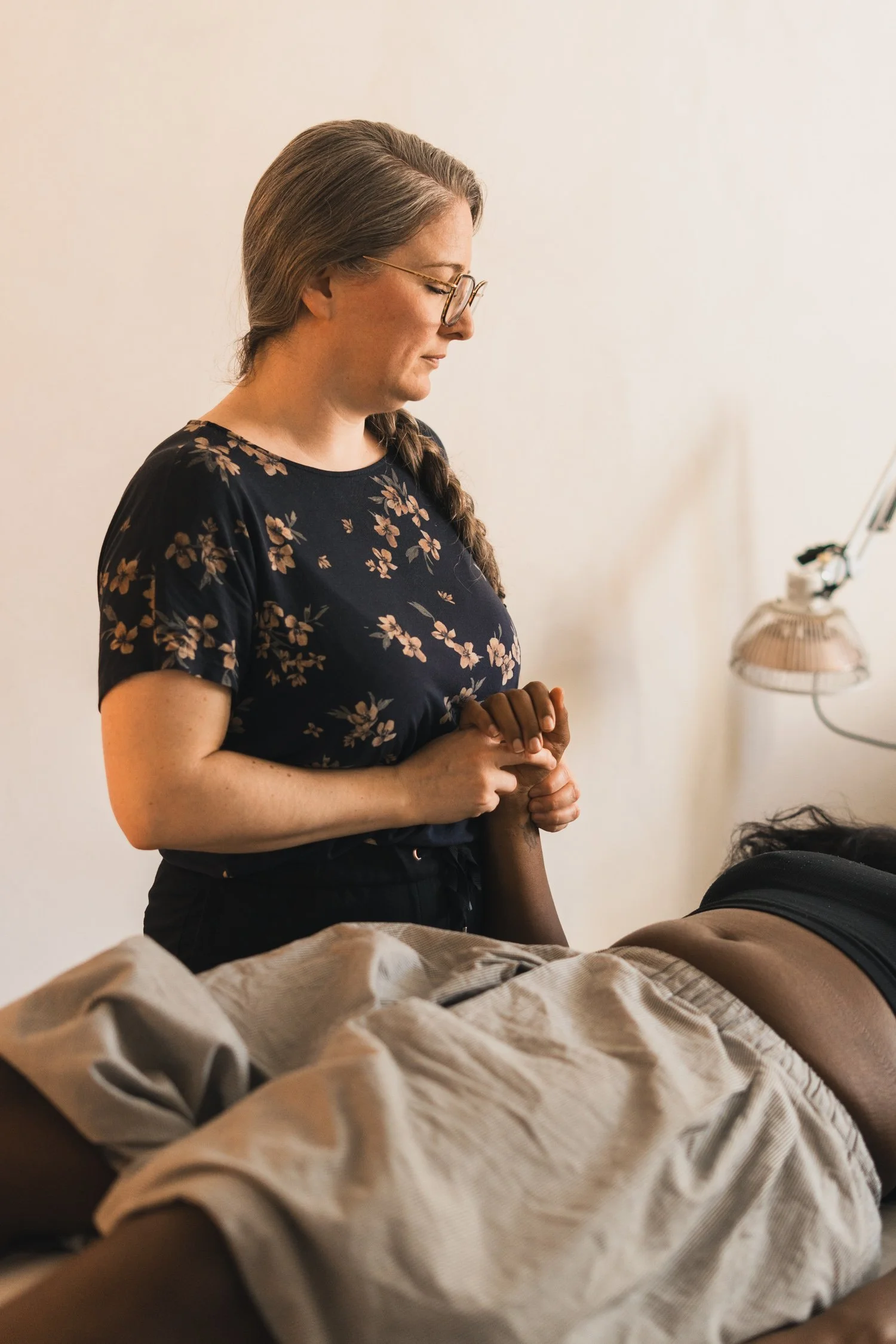 A woman with glasses and long hair in a braid is standing with her hands clasped, looking down at a person lying on a bed, who has tags on their arm, in a room with a plain wall and a lamp.