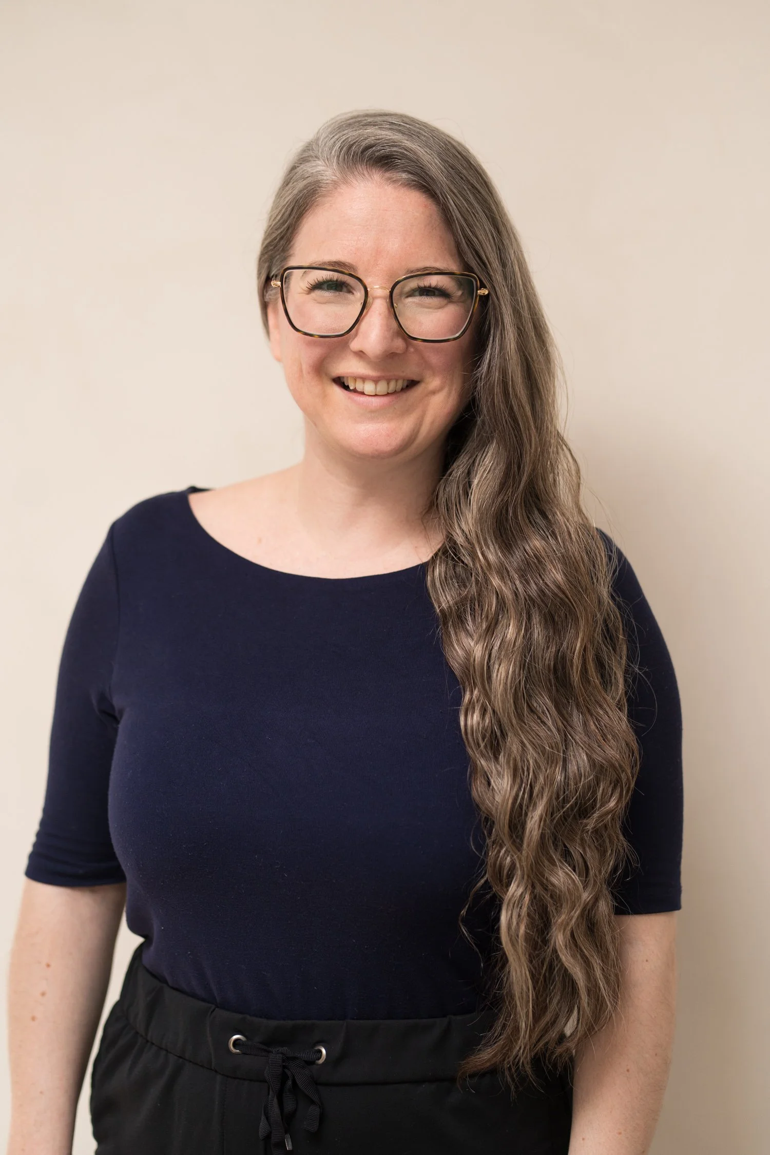 A woman with long, wavy brown hair, wearing glasses and a black top, smiling in front of a plain, light-colored wall.