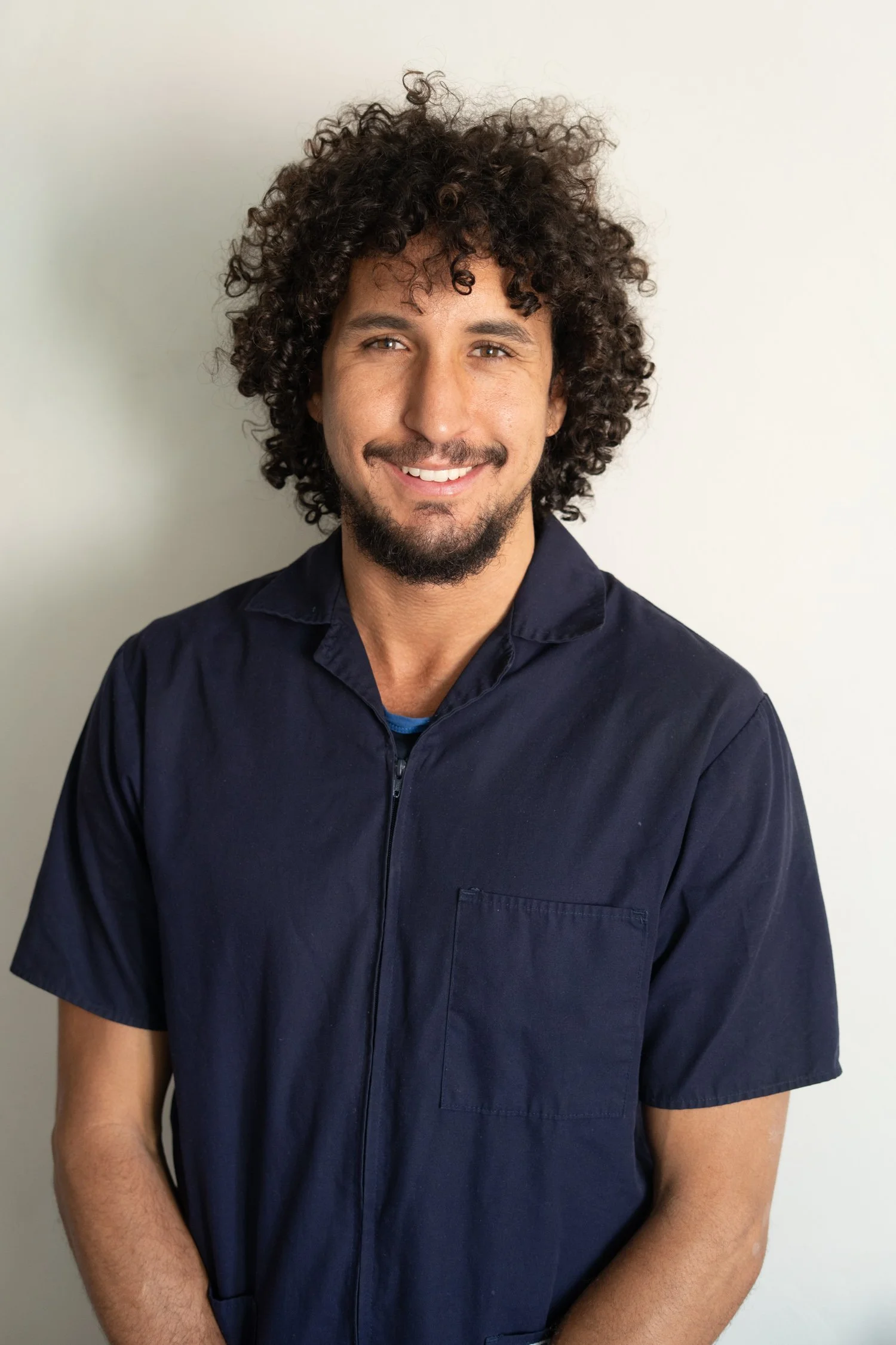A man with curly hair and a beard smiling, wearing a navy blue shirt, standing against a plain white wall.