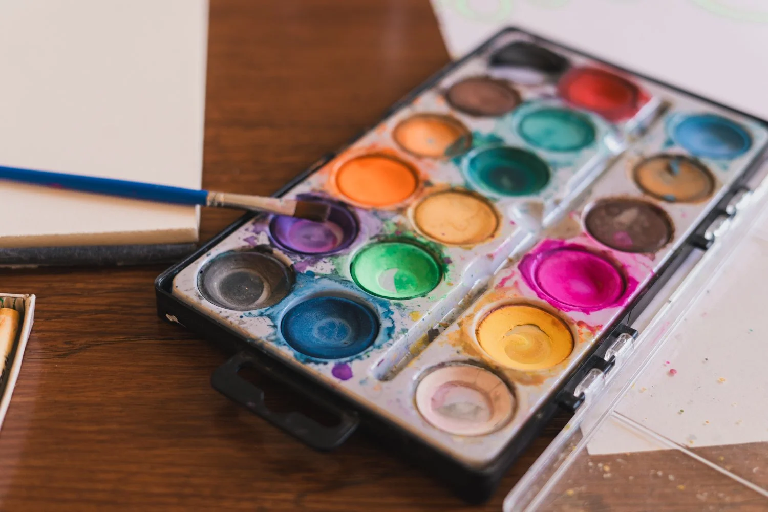 A watercolor paint set with various colors on a wooden table, a paintbrush resting in one of the colors, and a blank white sheet of paper nearby.
