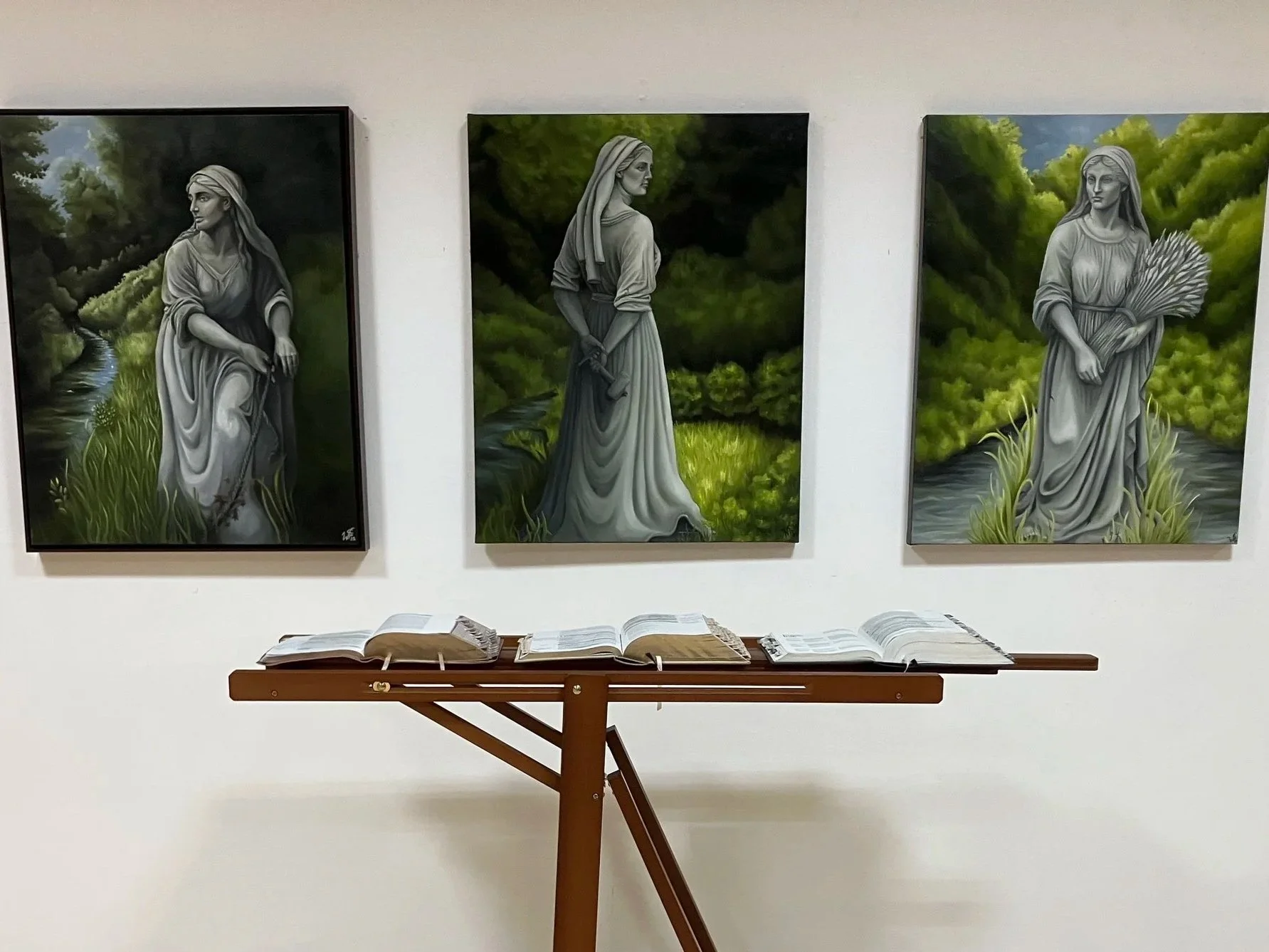 Three paintings of women with nature backgrounds displayed on a white wall, with open books on a wooden stand below.