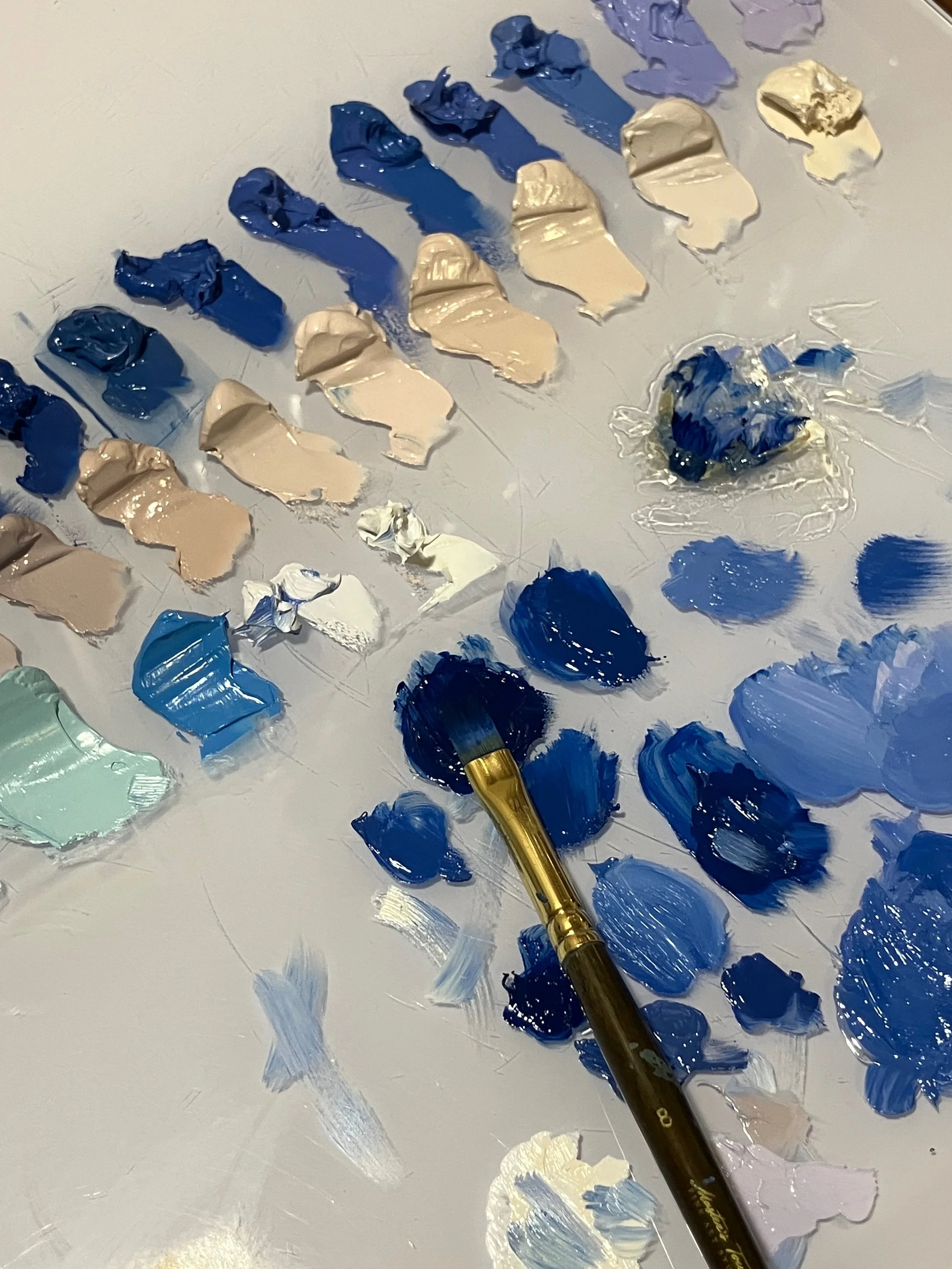 A paint palette with various shades of blue, beige, and white paint, along with a paintbrush resting on the palette, stained with blue paint.
