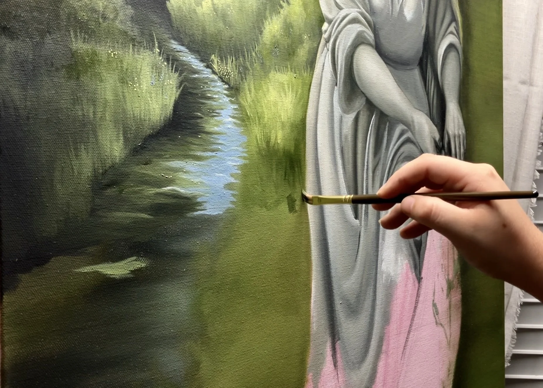 Close-up of an artist painting a realistic gray and white portrait of a woman sitting outdoors near a stream, with green grass and trees in the background.