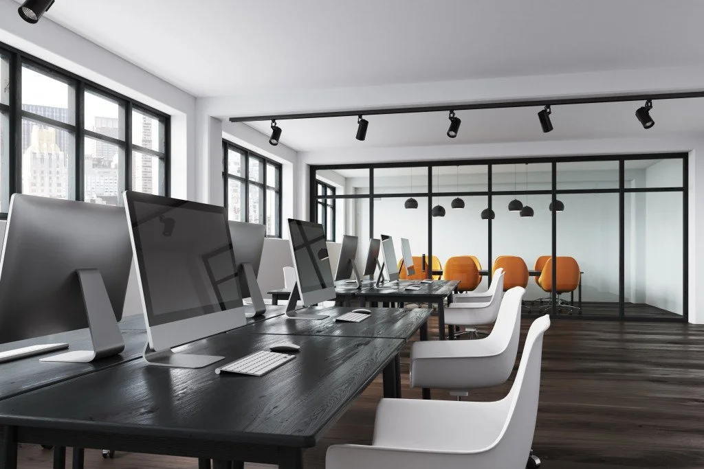 A modern office with multiple computer stations along a long desk, white chairs, large windows, and a glass enclosed meeting room with orange chairs inside. MSP covers PC/LapTops at $65 per device.